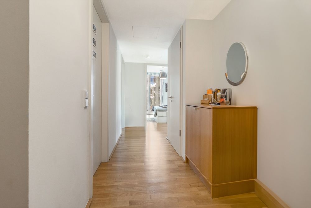 Heller Flur mit Holzfußboden, Kommode, rundem Spiegel und Blick in ein Zimmer.