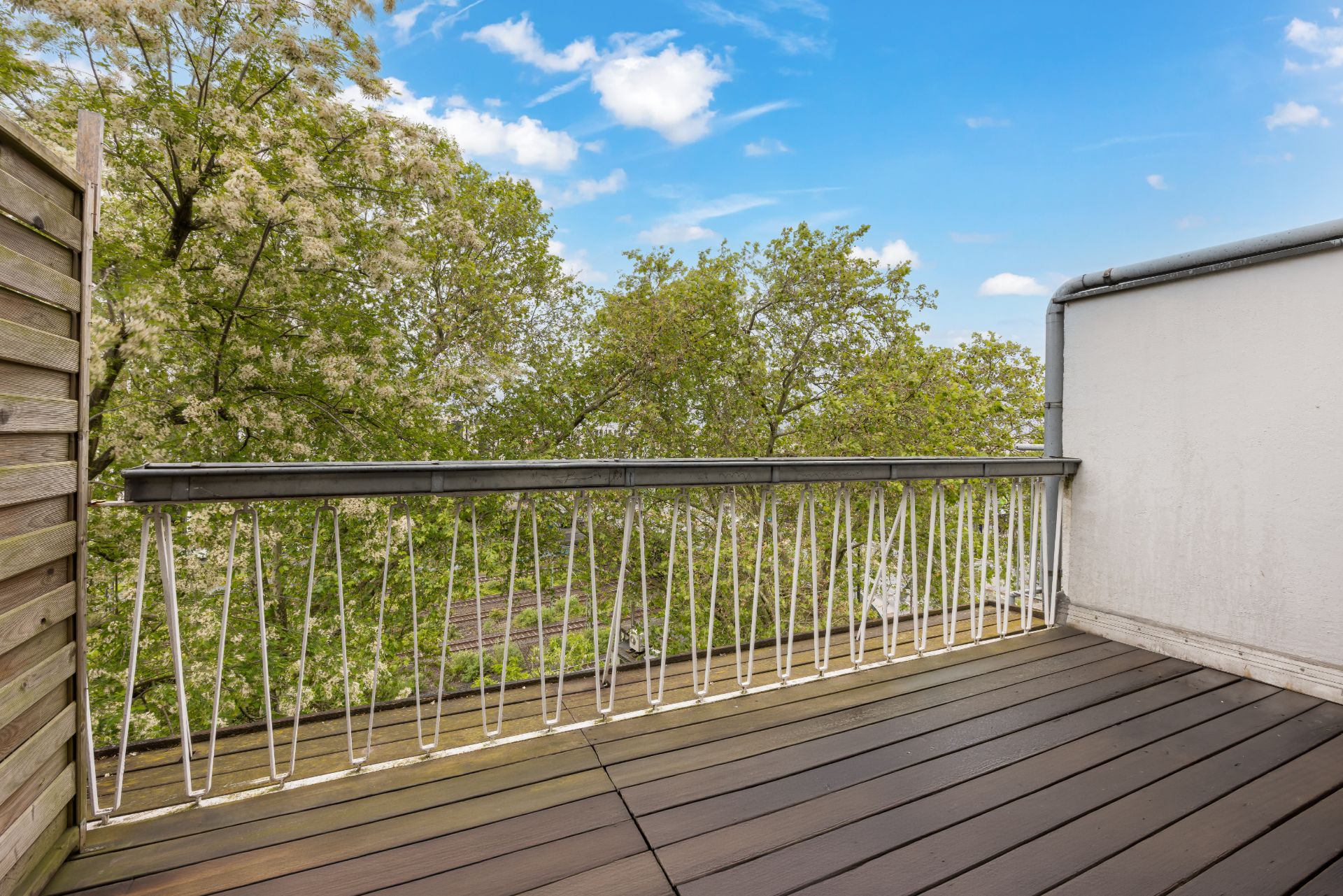 Balkon mit Holzboden, Geländer und Blick auf grüne Bäume bei blauem Himmel.