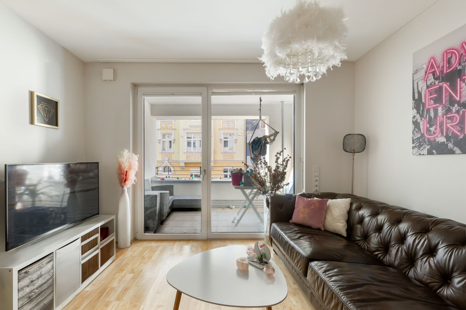 Helles Wohnzimmer mit Ledersofa, TV, Couchtisch und Zugang zu einem Balkon mit Sitzbereich.