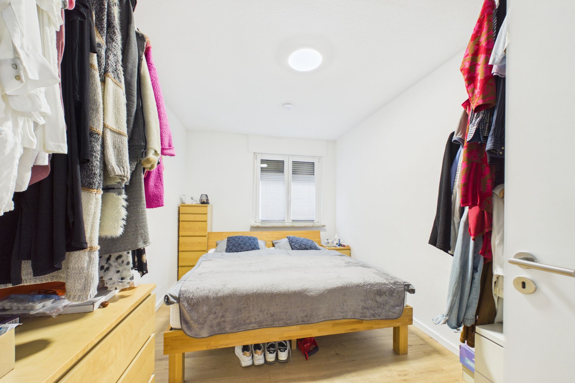 Schlafzimmer mit Doppelbett, Holzkommoden, Fenstern und Kleidung an den Seiten.