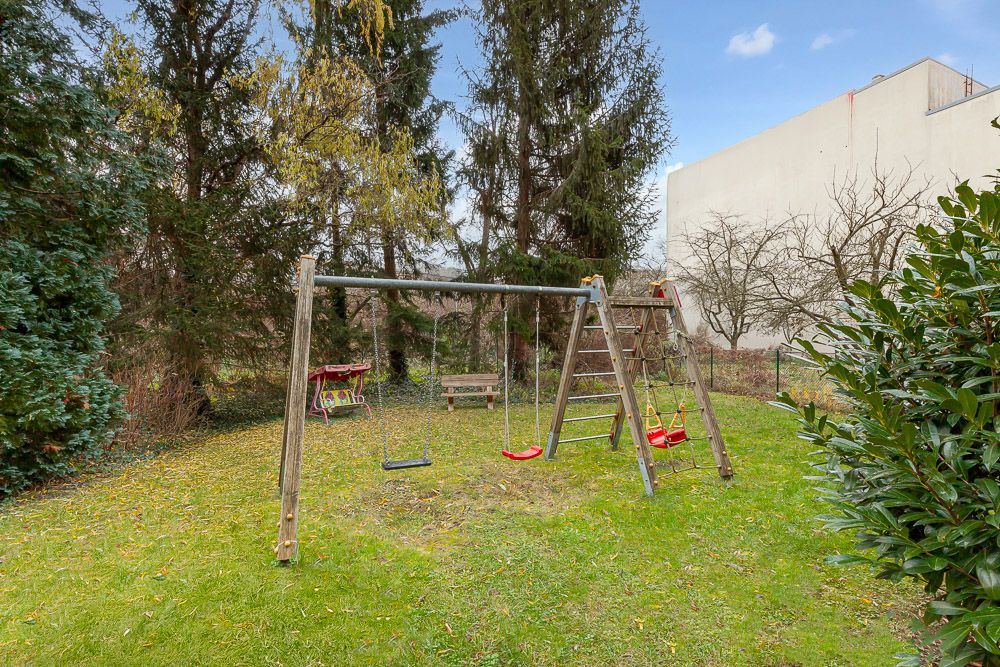 Garten mit Holzspielplatz, Schaukeln und Kletterleiter auf Rasen, umgeben von Bäumen.
