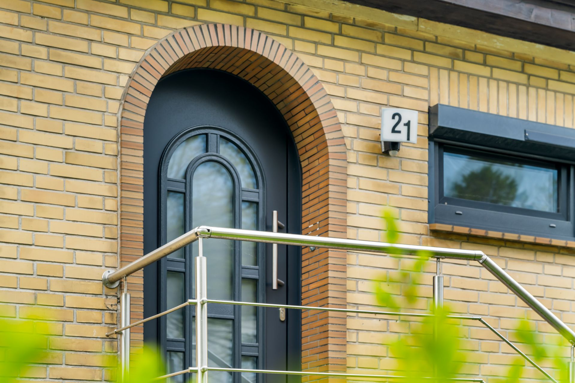 Schwarze Haustür mit Rundbogen, Hausnummer 21, daneben ein Fenster und ein Metallgeländer.