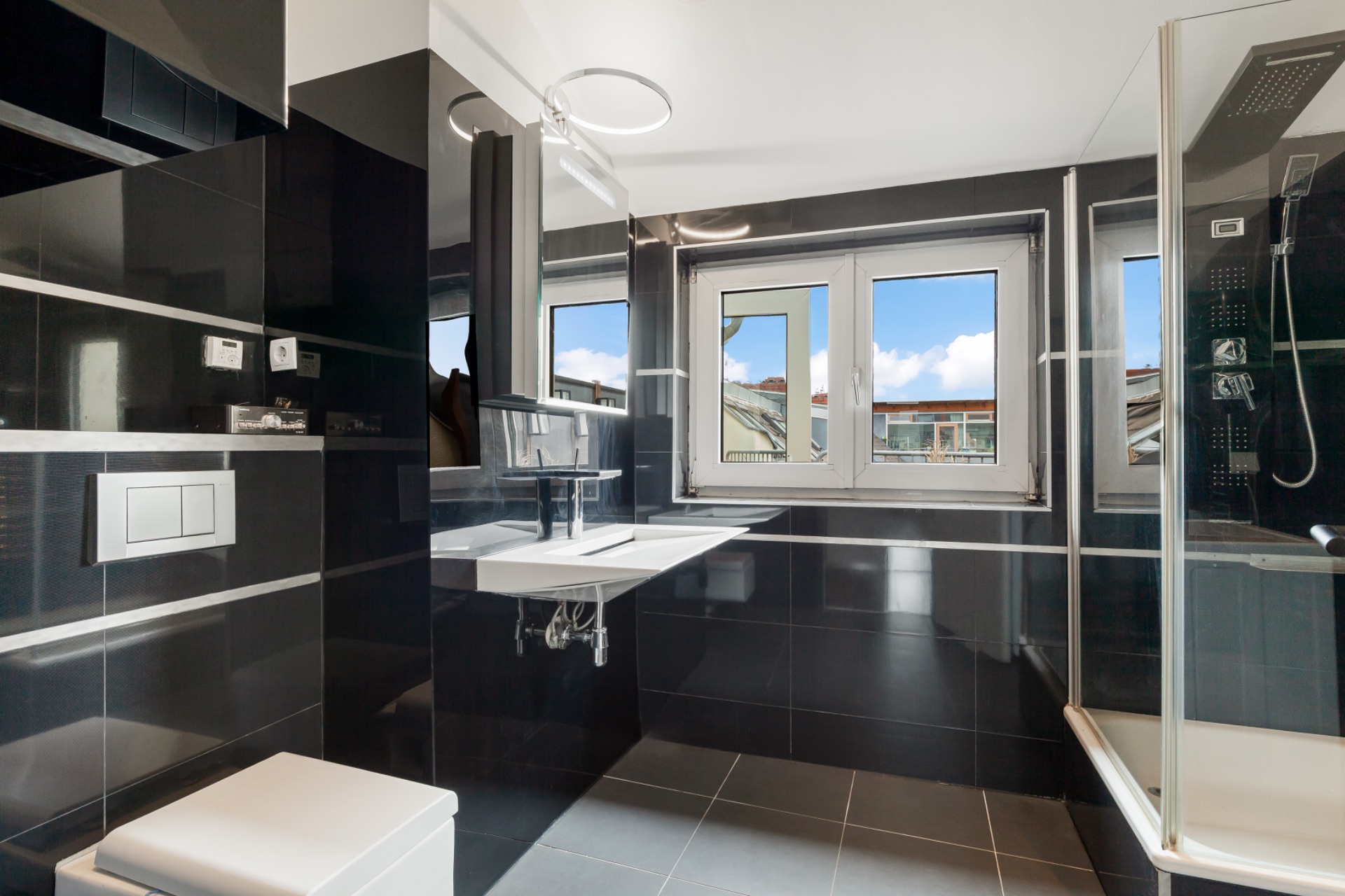 Modernes Badezimmer mit schwarzer Fliesenwand, Dusche, Fenster, Waschbecken und Toilette.