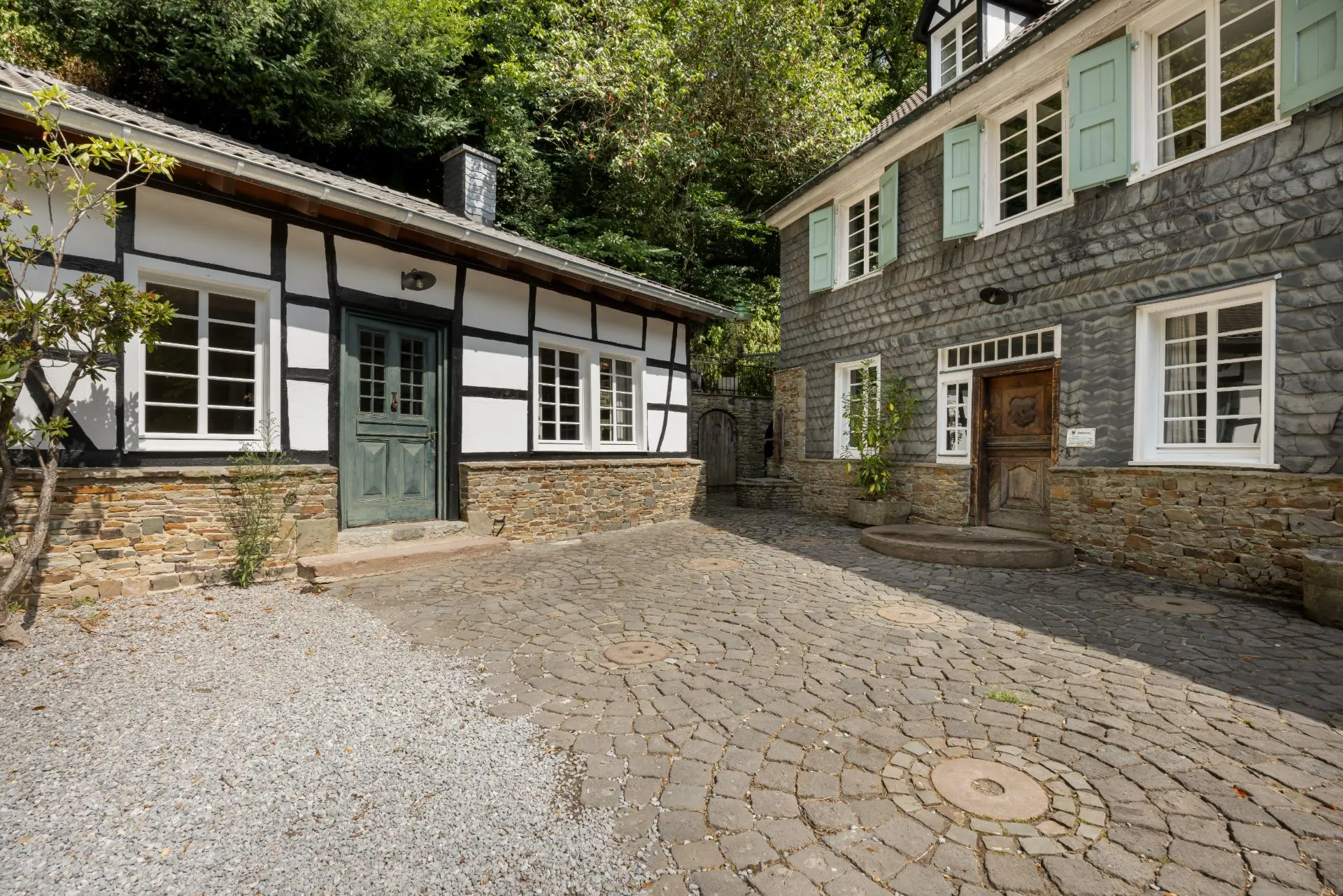 Gepflasterter Hof mit zwei traditionellen Häusern, Fachwerk und Graustein, grüne Fensterläden.