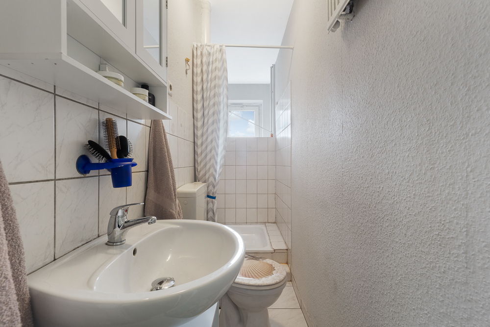 Schmales Badezimmer mit Waschbecken, Toilette, Dusche und Wandregal.