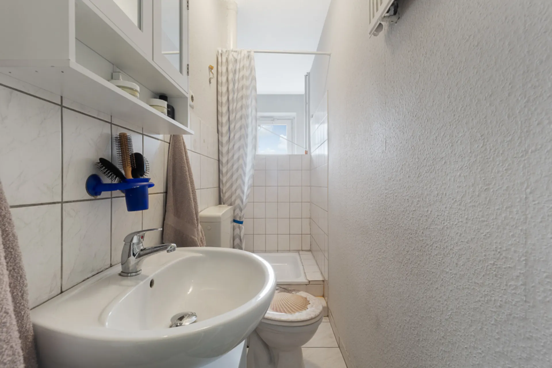 Schmales Badezimmer mit Waschbecken, Toilette, Dusche und Wandregal.