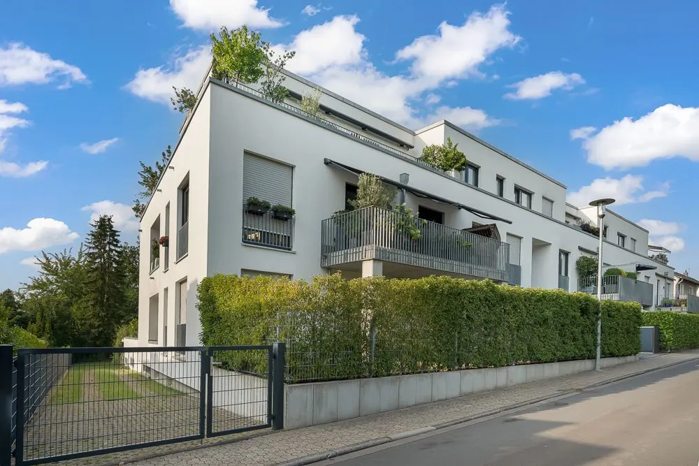 Modernes Wohngebäude mit Balkonen, Grünpflanzen und Zaun an einer ruhigen Straße.