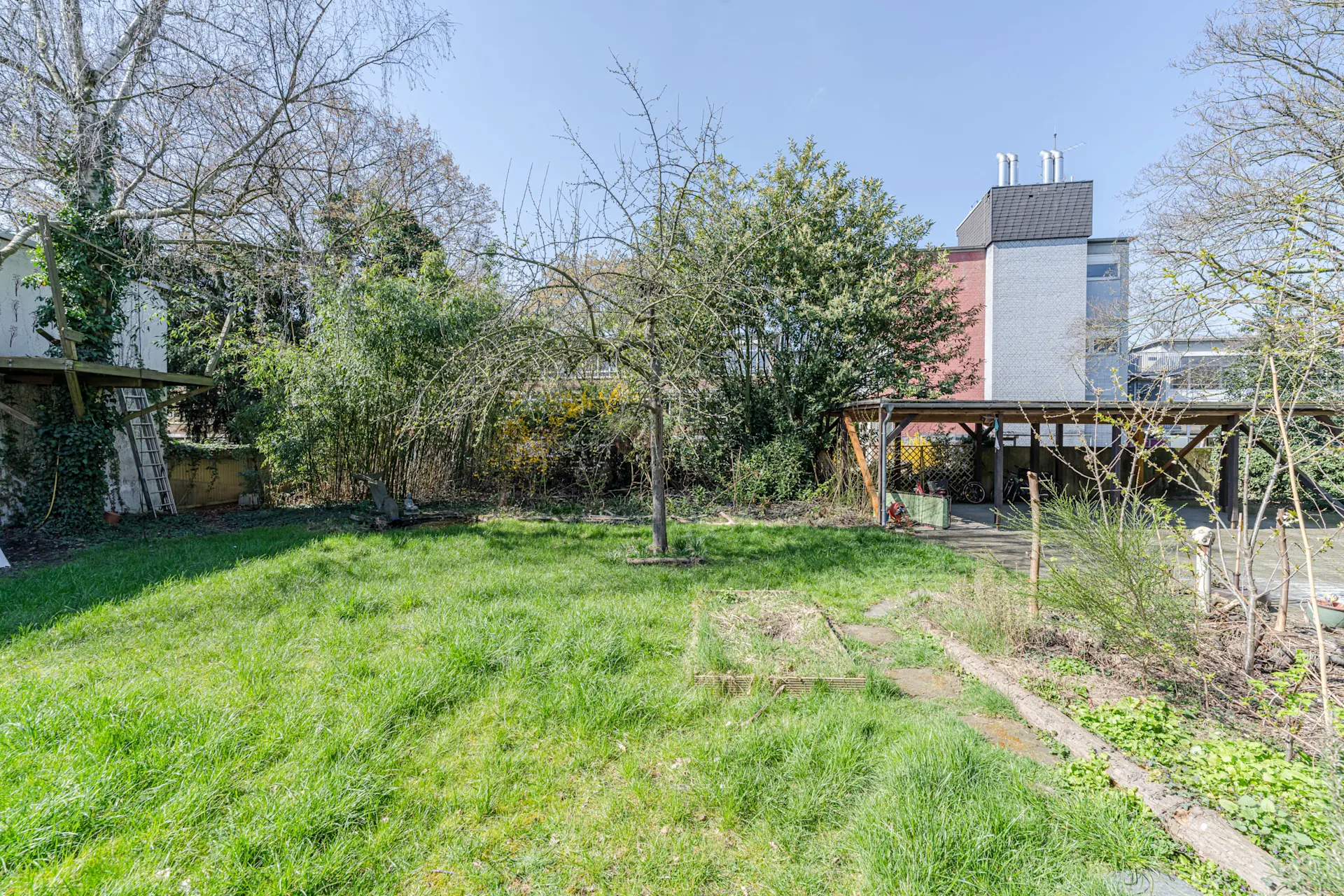 Grüner Garten mit einem Baum, einem Beet, Rasenfläche und einem Carport im Hintergrund.