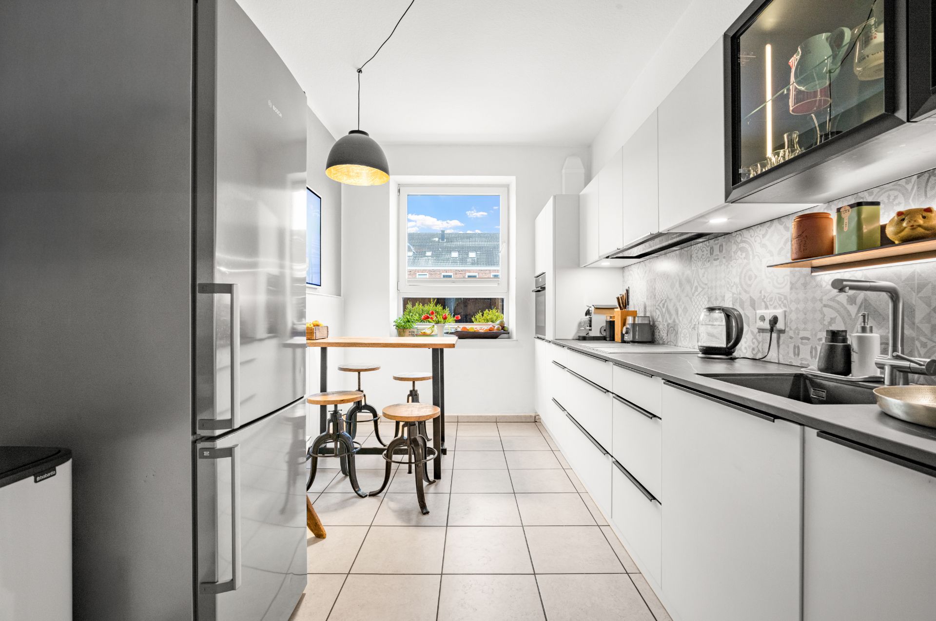 Moderne, helle Küche mit weißen Schränken, großem Kühlschrank und Esstisch am Fenster.