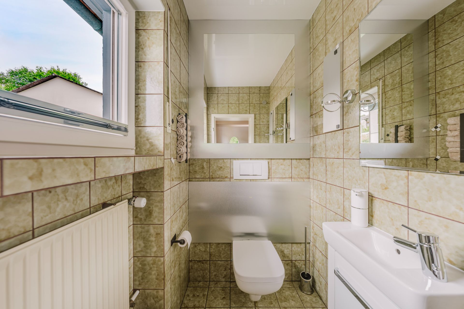 Kleines Badezimmer mit Wandtoilette, Waschbecken, Spiegel und geöffnetem Fenster.