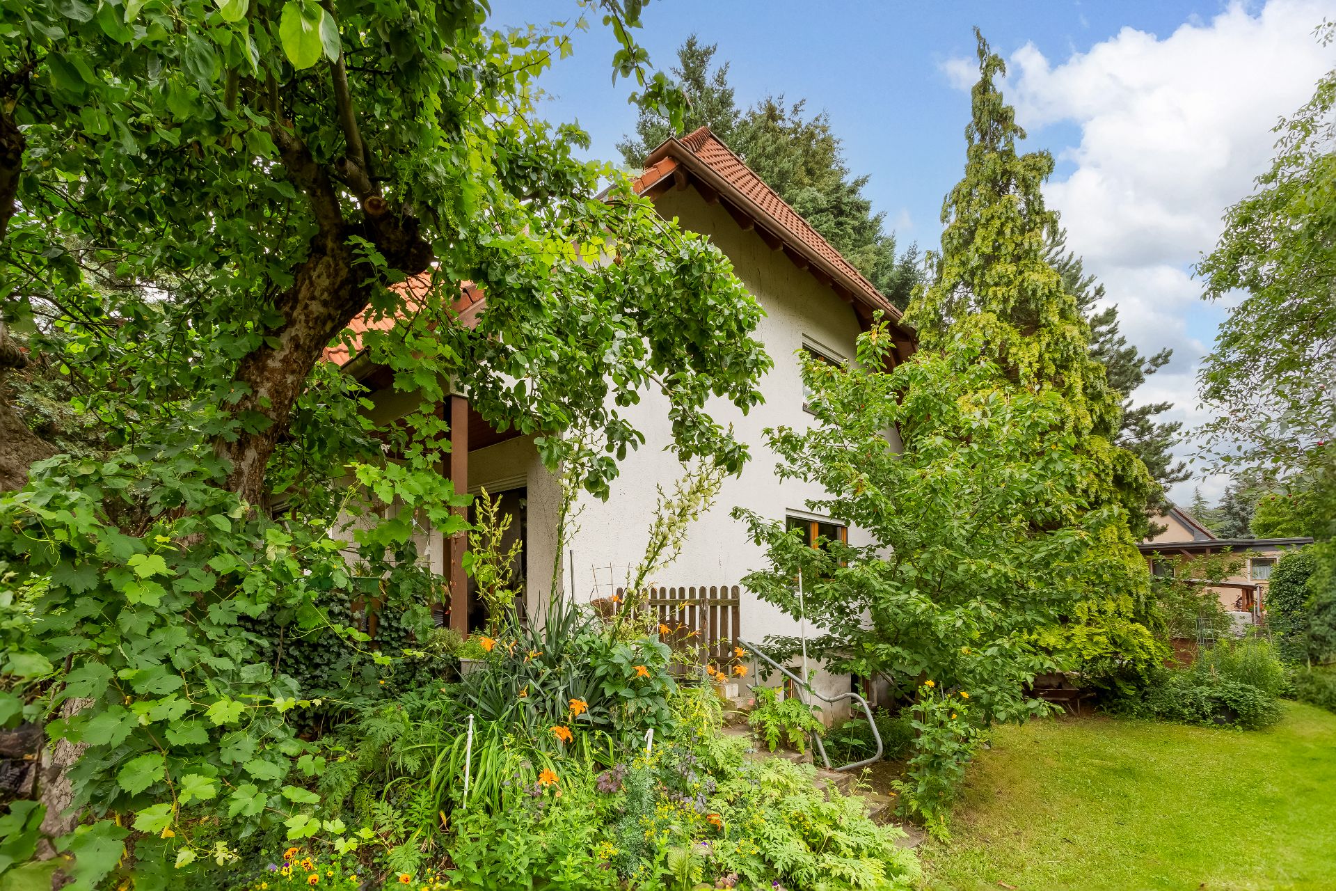 Einfamilienhaus mit rotem Dach, umgeben von dichtem grünen Garten und verschiedenen Bäumen.