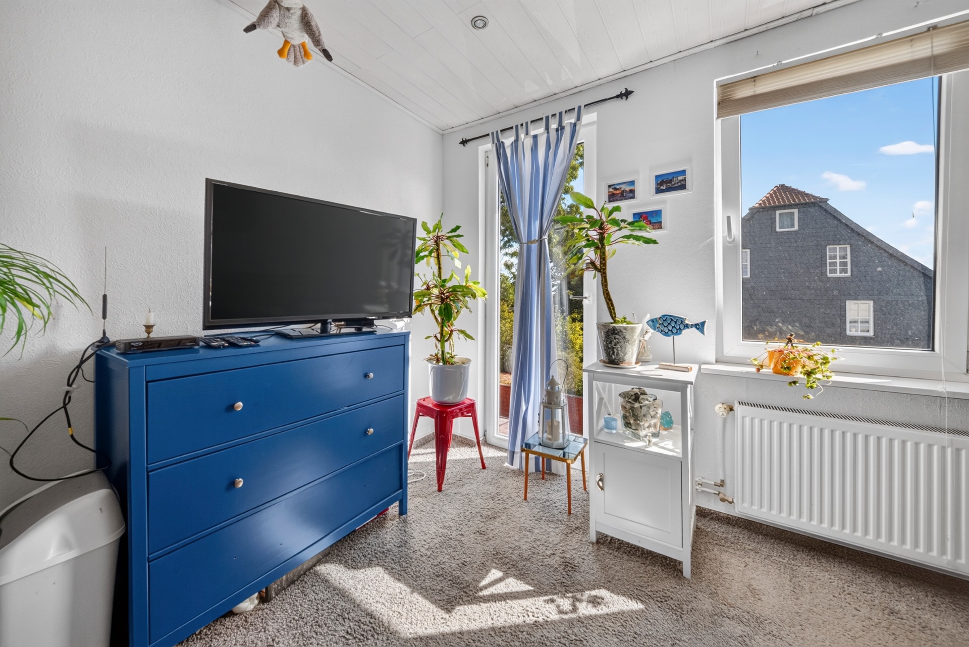 Helles Zimmer mit blauem Sideboard, Fernseher, Pflanzen und großem Fenster.