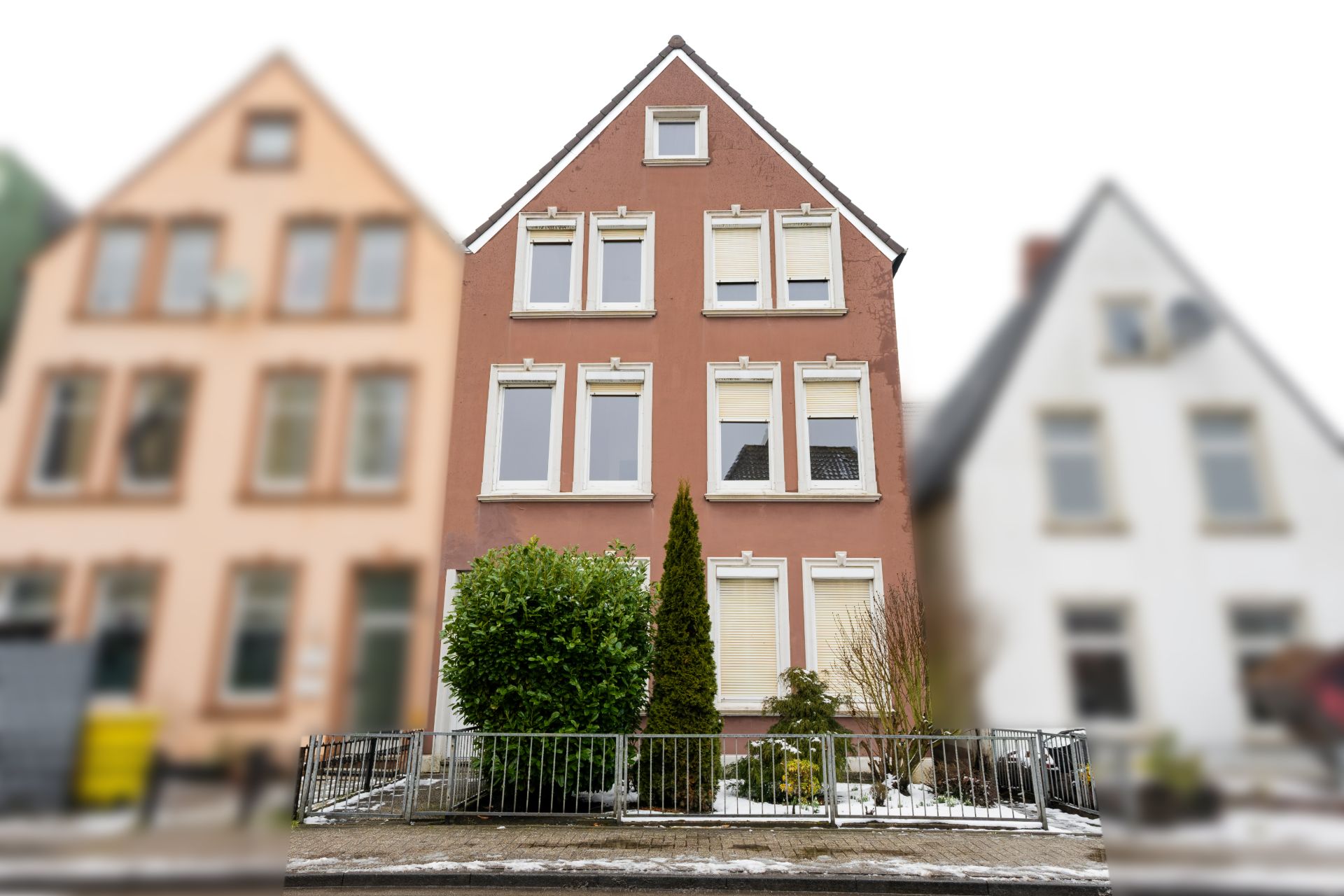 Dreistöckiges Wohnhaus mit spitzem Dach, Vorgarten und Zaun an der Straße.