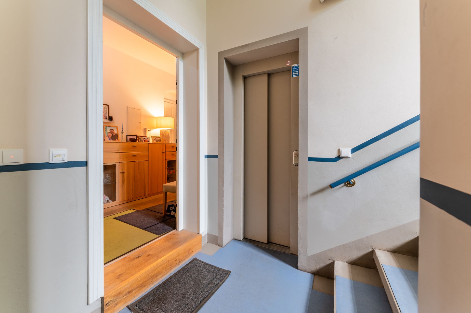Hausflur mit Aufzug, Treppe und Blick in ein Wohnzimmer mit Möbeln und Lampe.