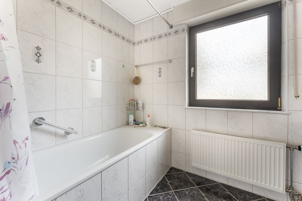 Helles Badezimmer mit Badewanne, Fenster und Heizung, gefliest mit weißen Fliesen.