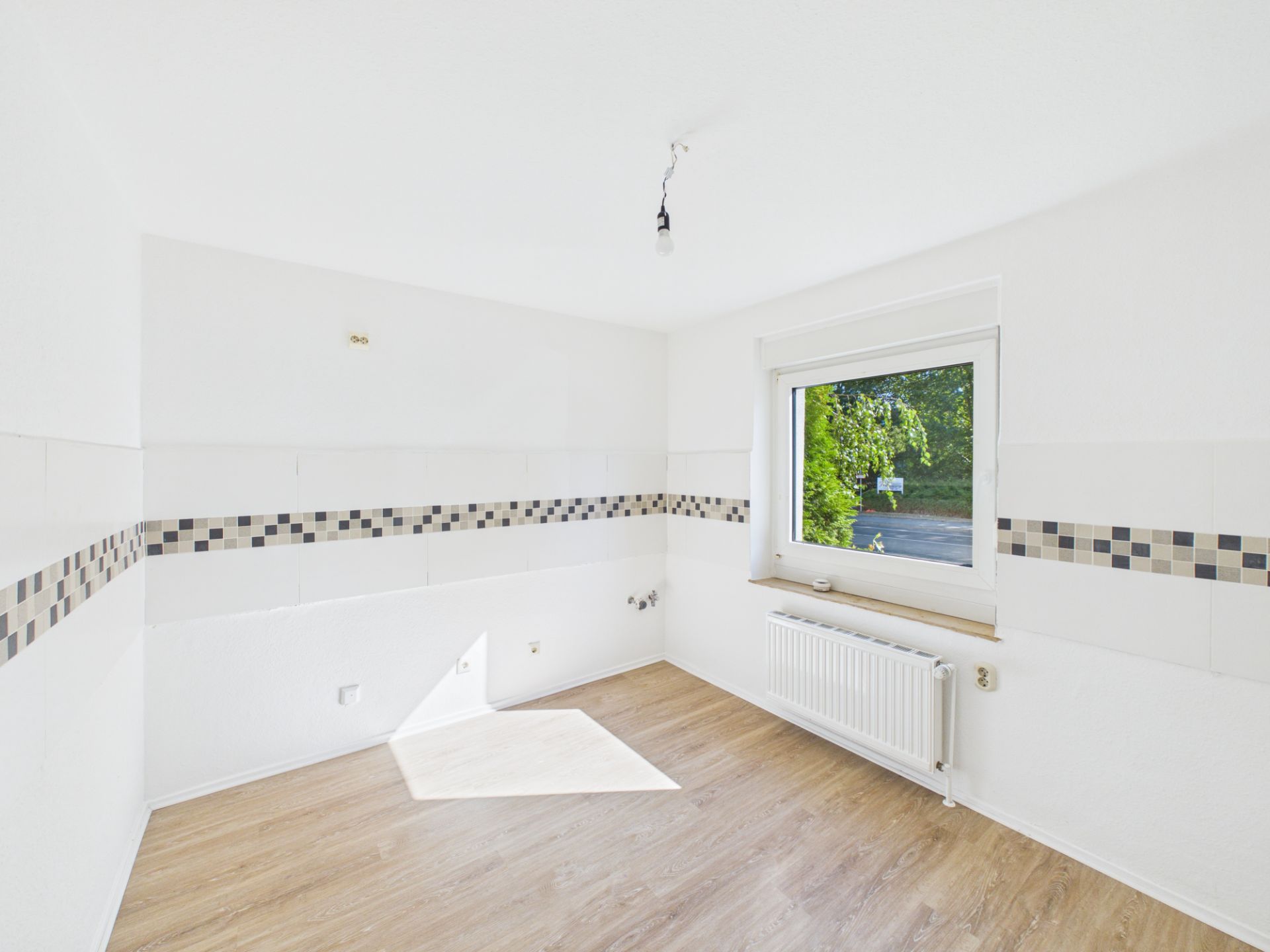 Leerer, heller Raum mit Fliesenbordüre, Fenster und Heizkörper, Holzboden.