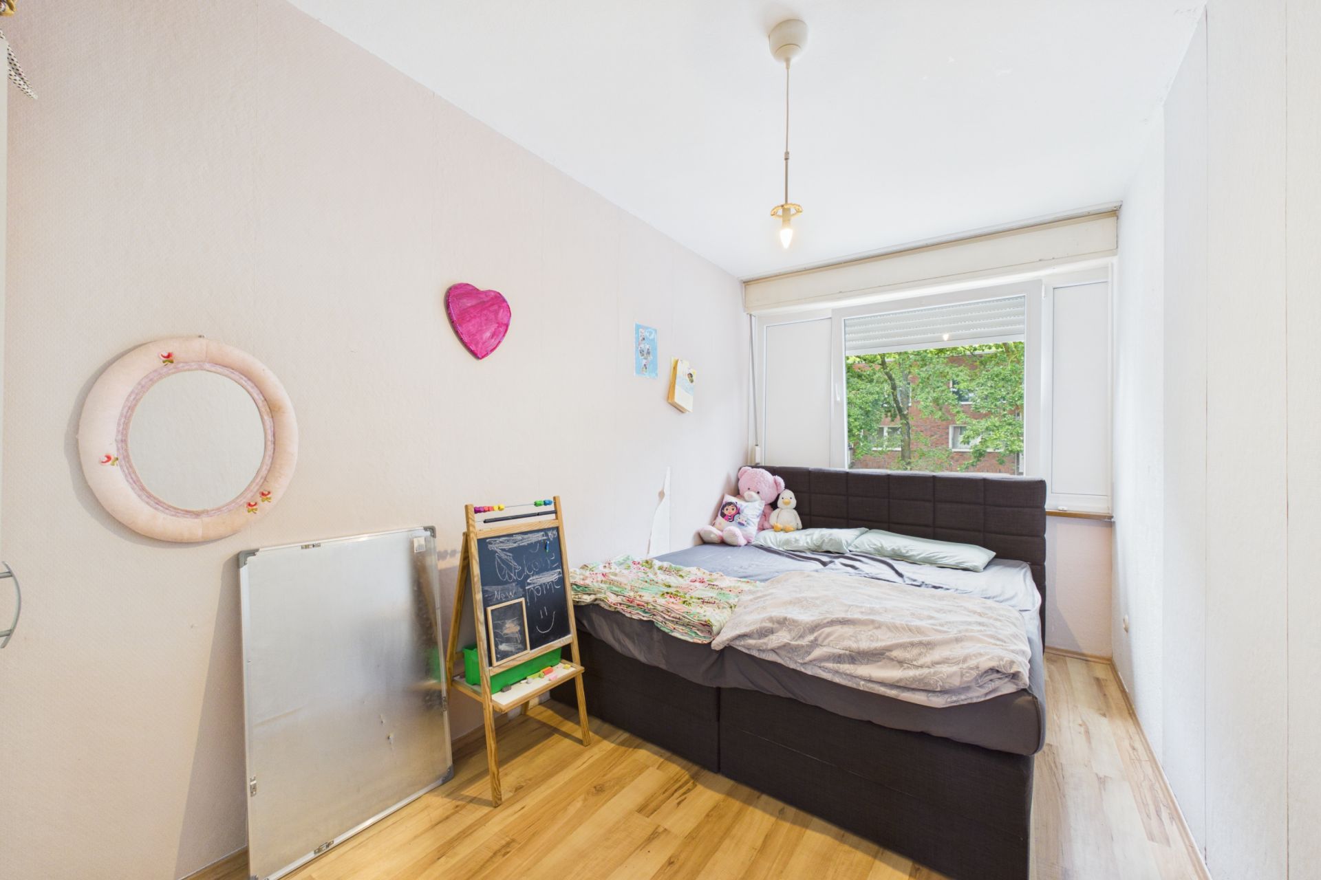 Helles Schlafzimmer mit Bett, Tafel, Spiegel und Kuscheltieren am Fenster.