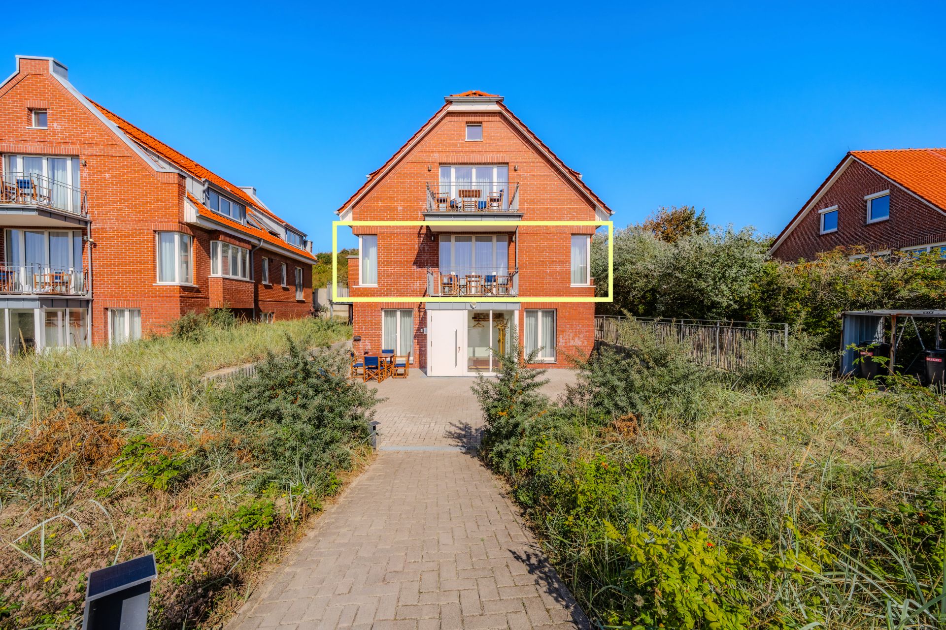 Ein rotes Backsteinhaus mit Balkon und zwei Etagen, umgeben von Grün und Gehweg.
