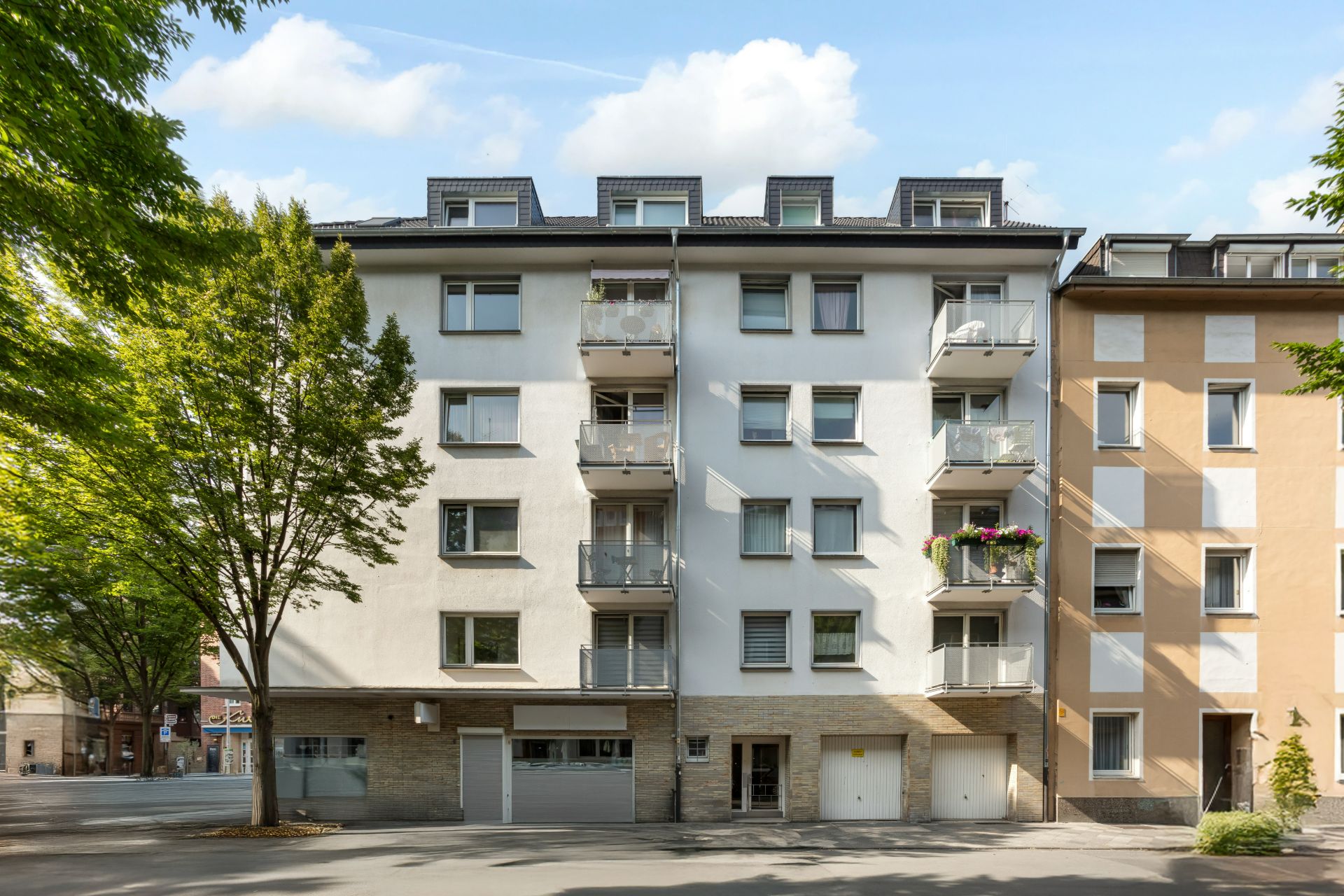 Mehrfamilienhaus mit Balkonen, fünf Stockwerke, Bäume an der Straße, helle Fassade.
