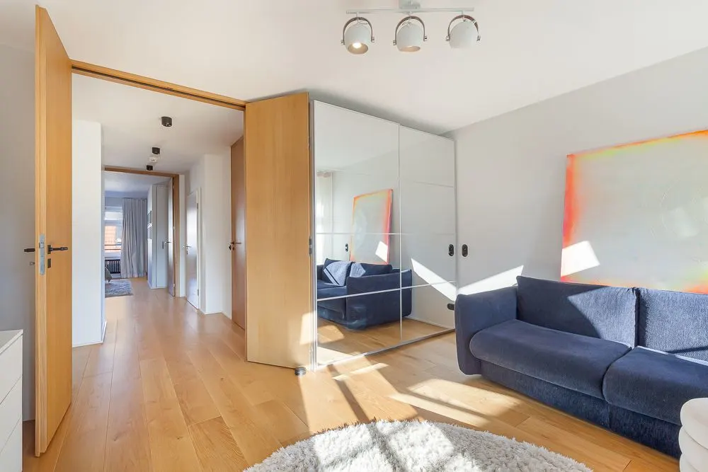 Helles Zimmer mit Holzfußboden, Spiegelkleiderschrank, blauer Couch und Teppich.