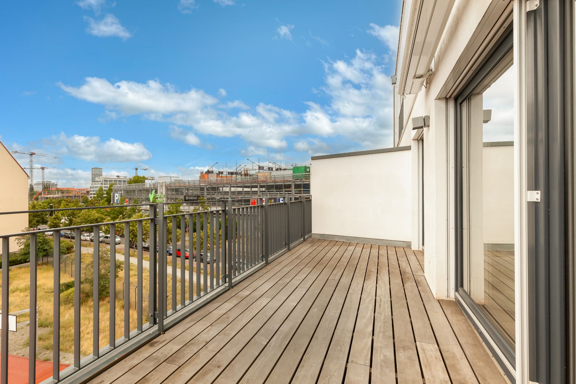 Geräumiger Balkon mit Holzboden, Metallgeländer und Blick auf die Stadt bei blauem Himmel.