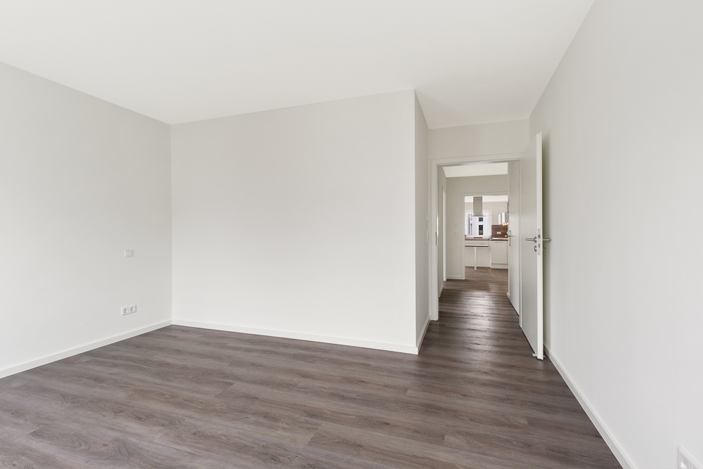 Leeres Zimmer mit weißen Wänden und dunklem Holzboden, Tür führt in einen Flur.