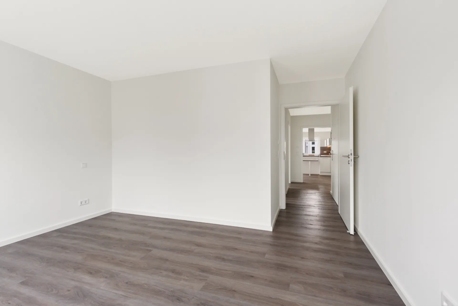 Leeres Zimmer mit weißen Wänden und dunklem Holzboden, Tür führt in einen Flur.