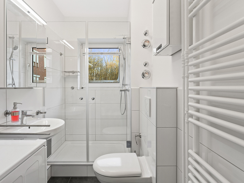 Modernes Badezimmer mit Dusche, WC, Waschbecken und Fenster, alles in weißen Fliesen.