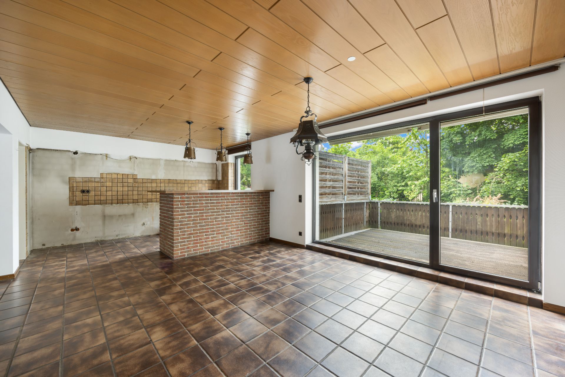 Leerer Wohnraum mit Fliesenboden, Holzdecke, Theke und großer Glastür zur Terrasse mit Blick ins Grüne.