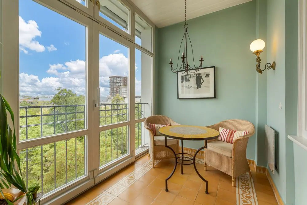 Heller Balkon mit zwei Korbstühlen, rundem Tisch, großer Fensterfront und Blick auf grüne Bäume.