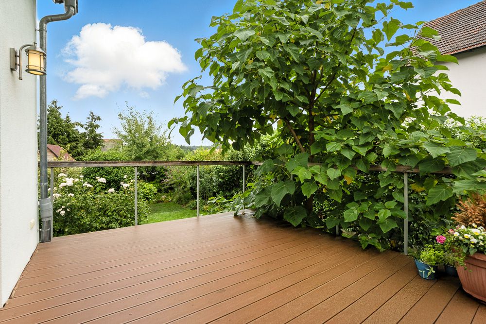 Holzterrasse mit Geländer, großen grünen Pflanzen und Blick auf einen Garten.