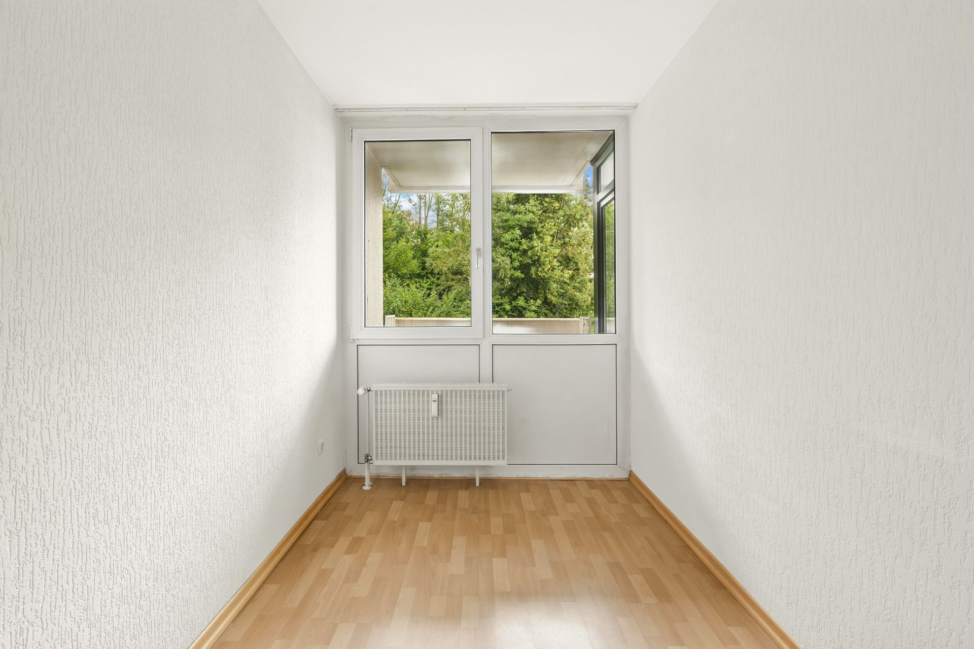 Leerer, schmaler Raum mit Holzfußboden, weiß gestrichenen Wänden und Fenster mit Blick ins Grüne.