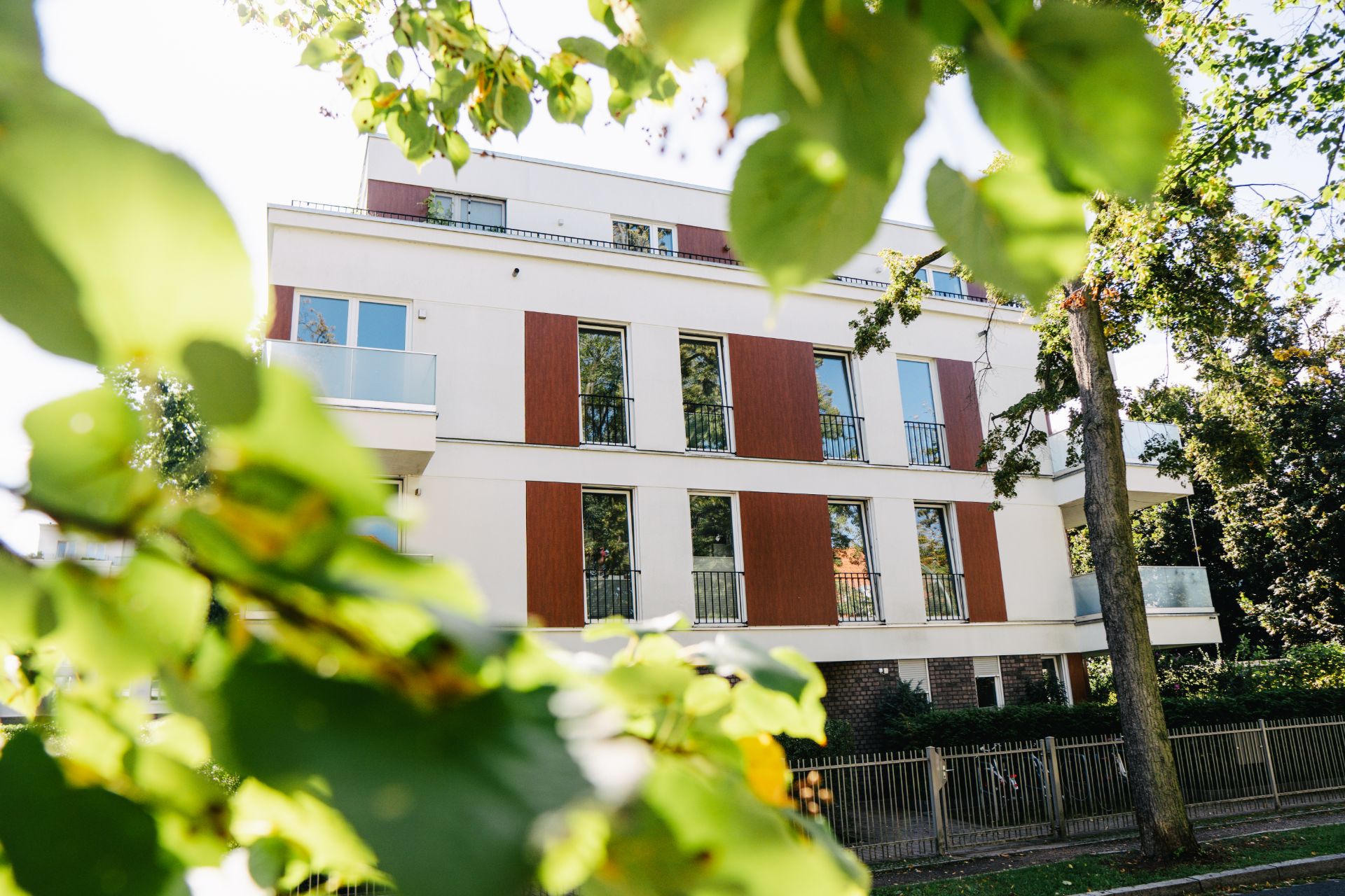 Modernes Wohnhaus mit Balkonen, Bäumen im Vordergrund und einem Zaun.