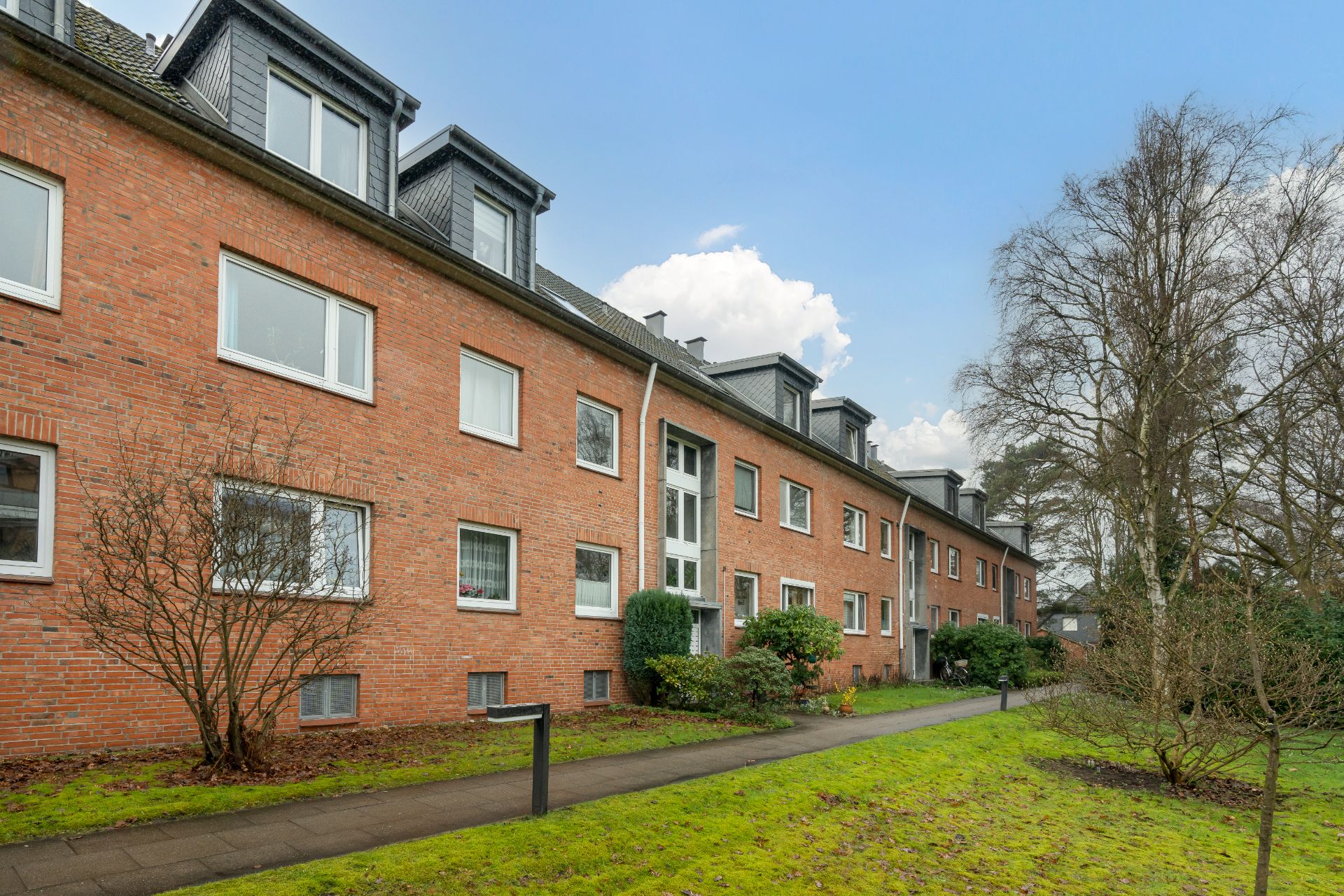 Mehrfamilienhaus mit roter Klinkerfassade, großen Fenstern und Vorgarten, Gehweg davor.