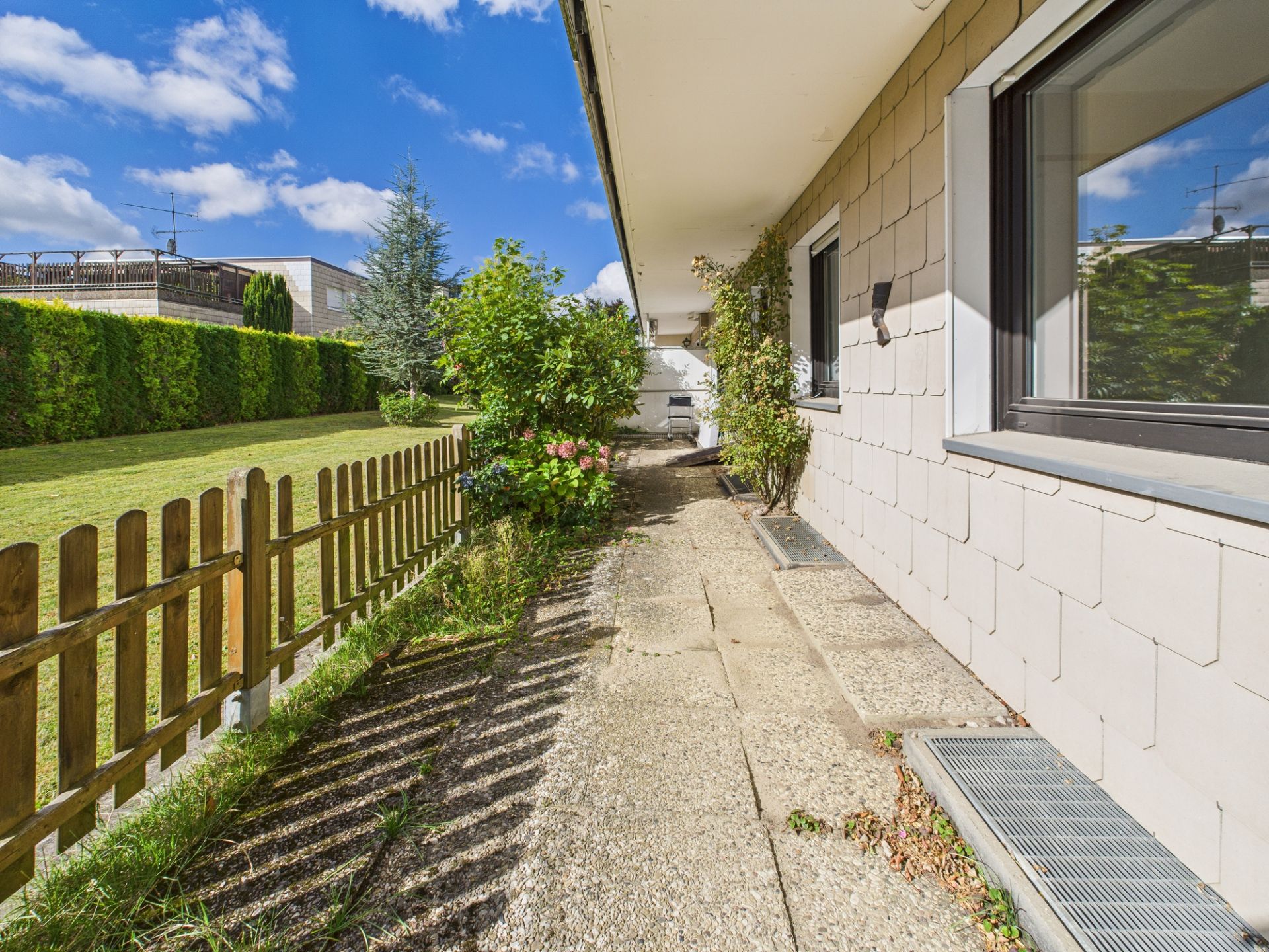Gepflasterter Weg entlang eines Hauses mit Holzzaun und Garten bei Sonnenschein.