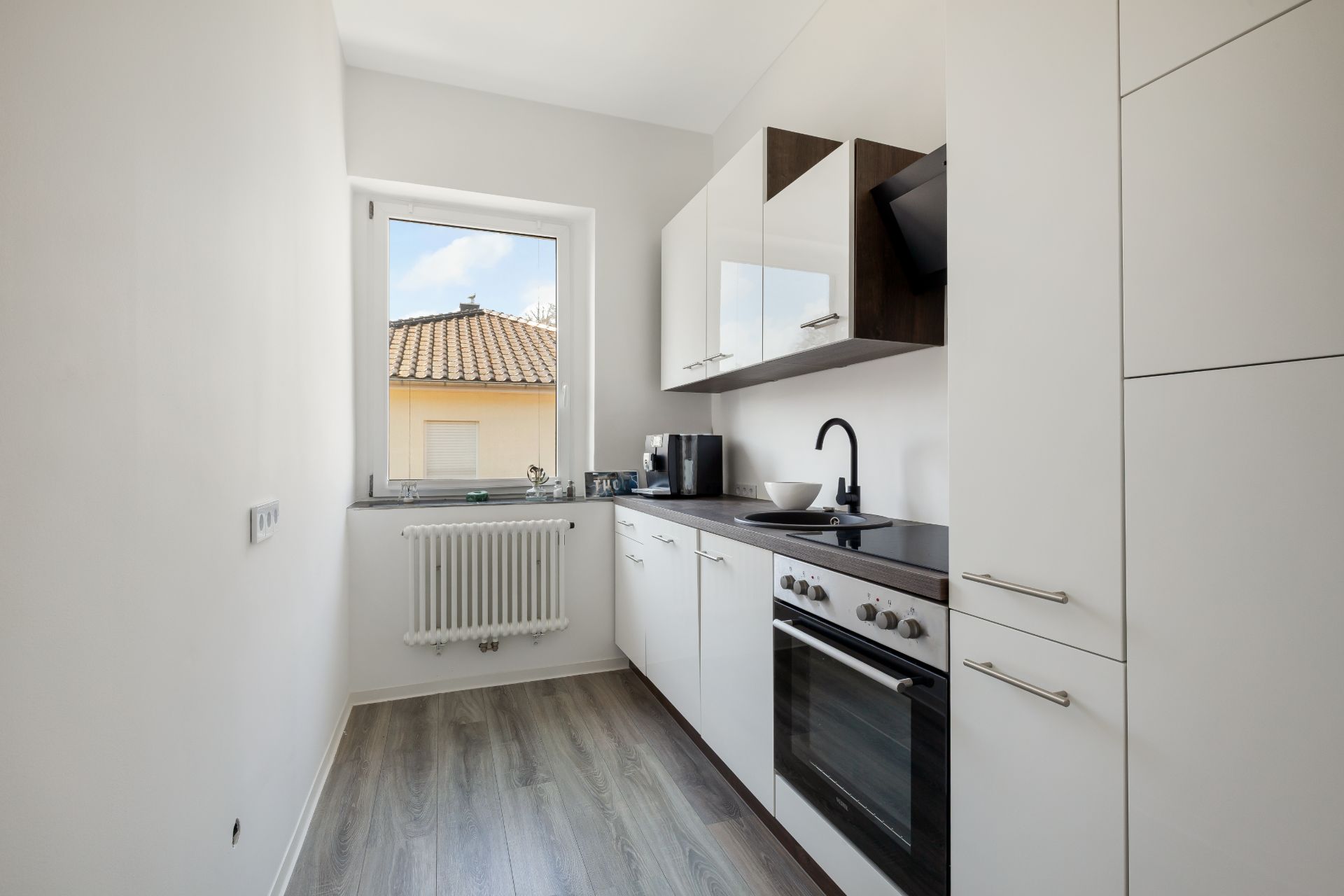 Moderne, schmale Küche mit weißen Schränken, Herd, Spüle und Fenster.