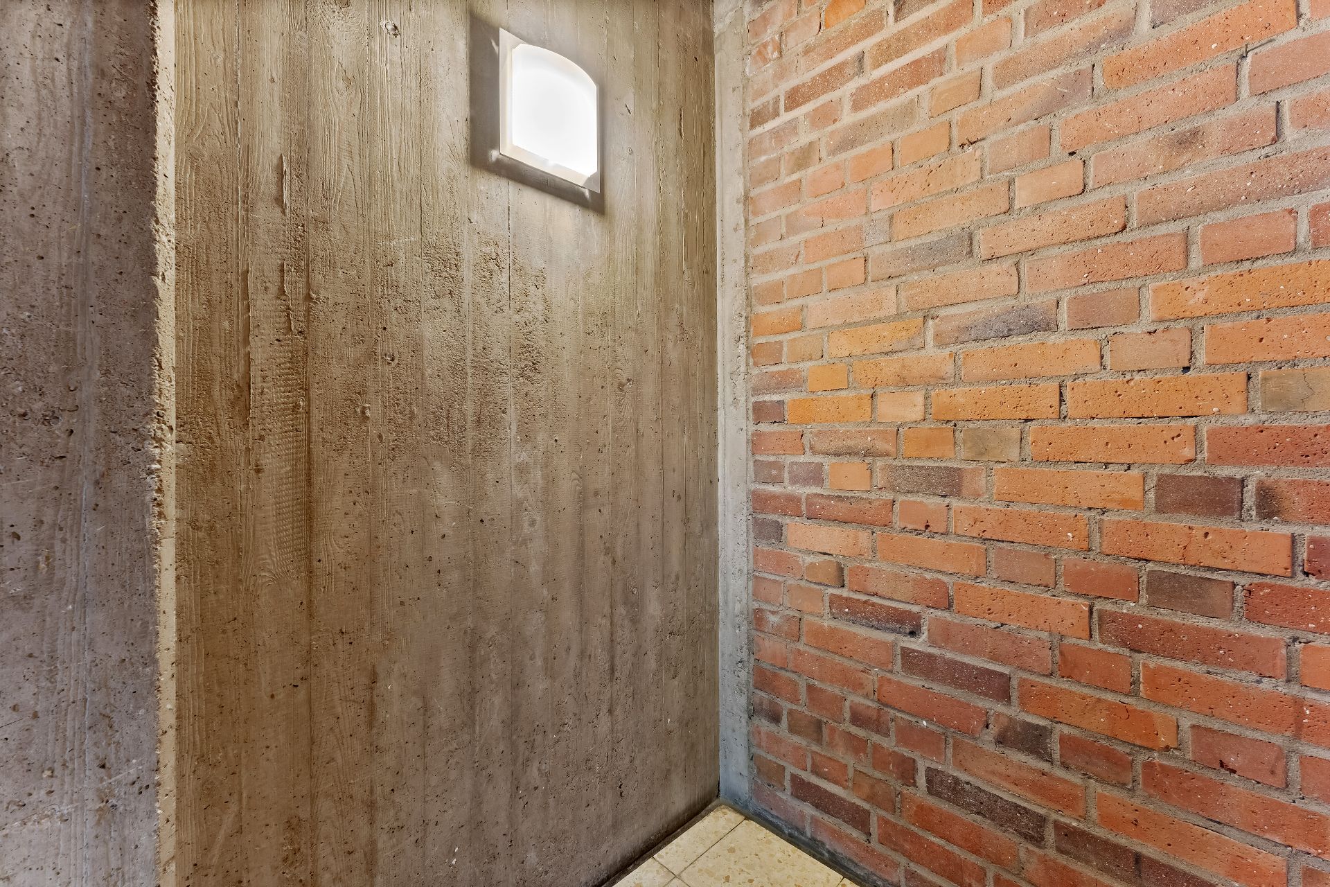 Ecke eines Raumes mit Beton- und Ziegelwand, Bodenfliesen und Wandlampe.