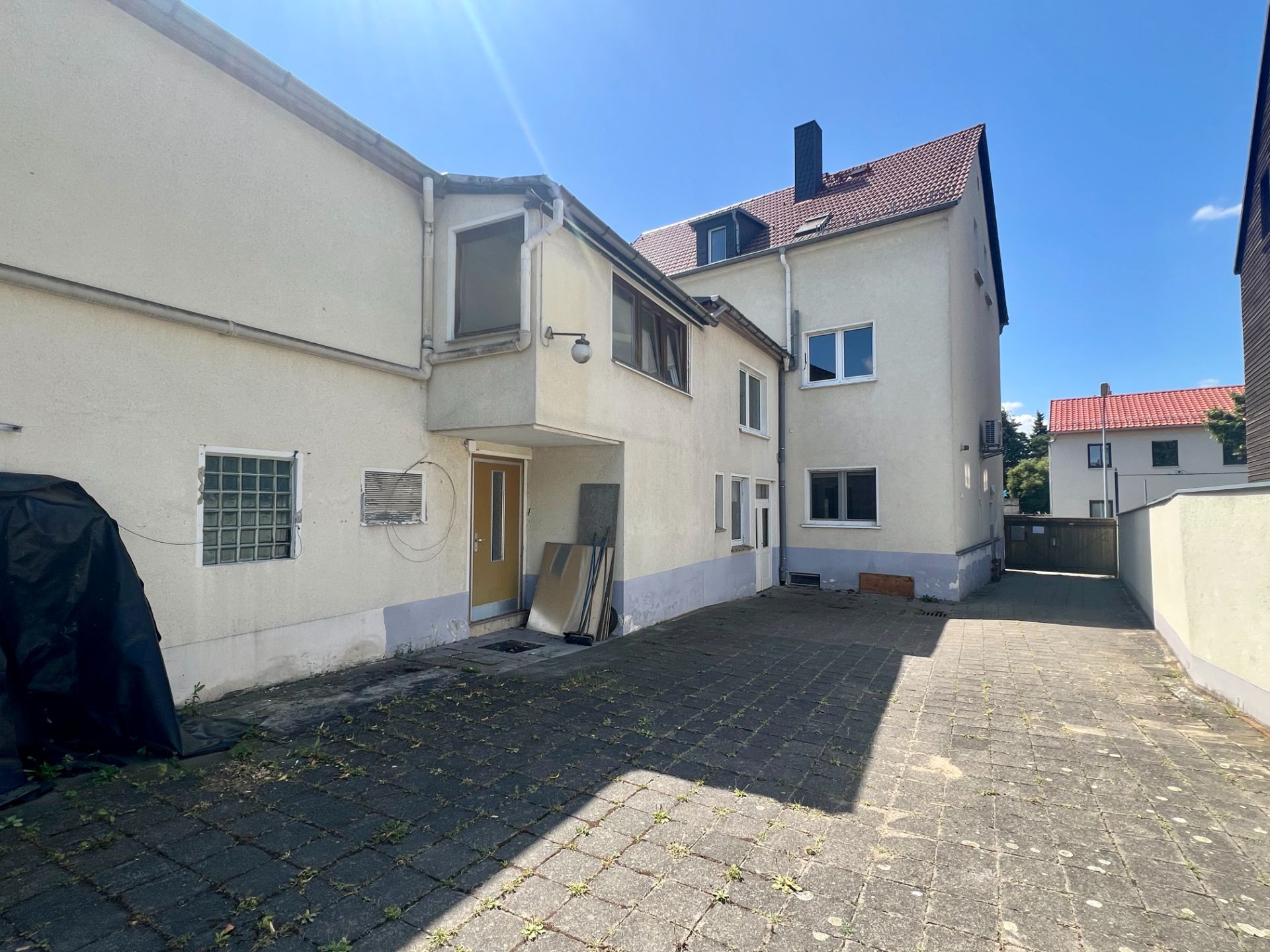 Gepflasterter Innenhof mit weißem Wohnhaus und Nebeneingang, blauer Himmel.