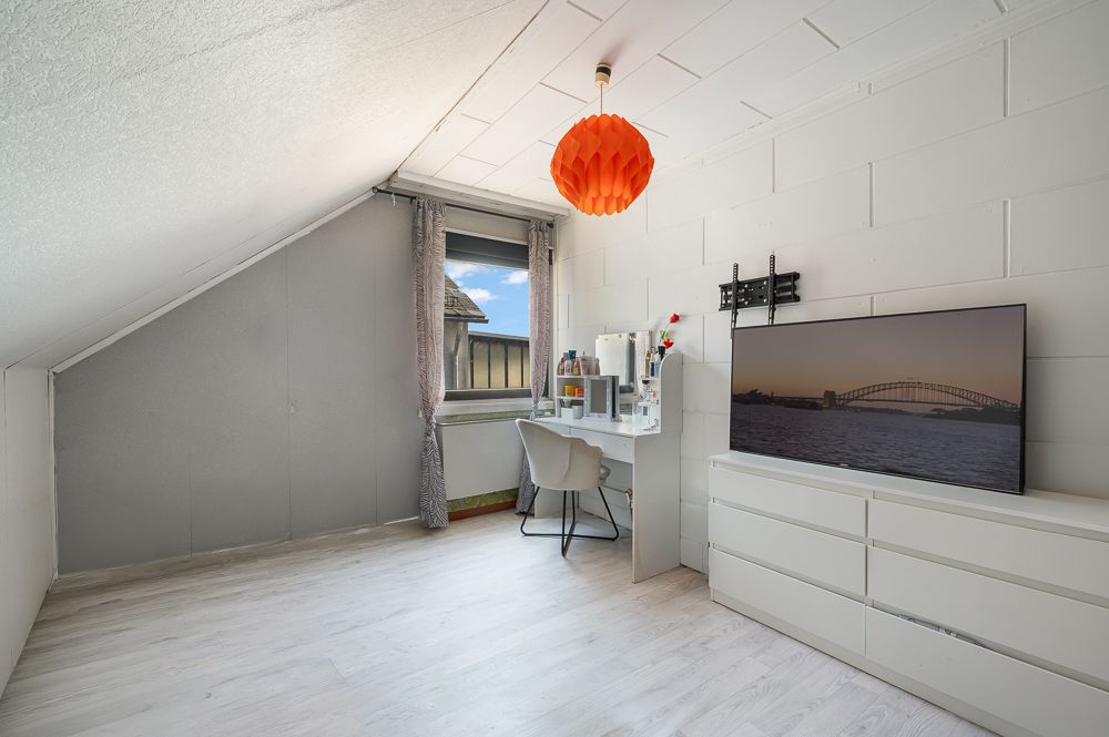 Helles Zimmer mit Schreibtisch, Stuhl, Kommode, Fernseher und orangener Deckenlampe.