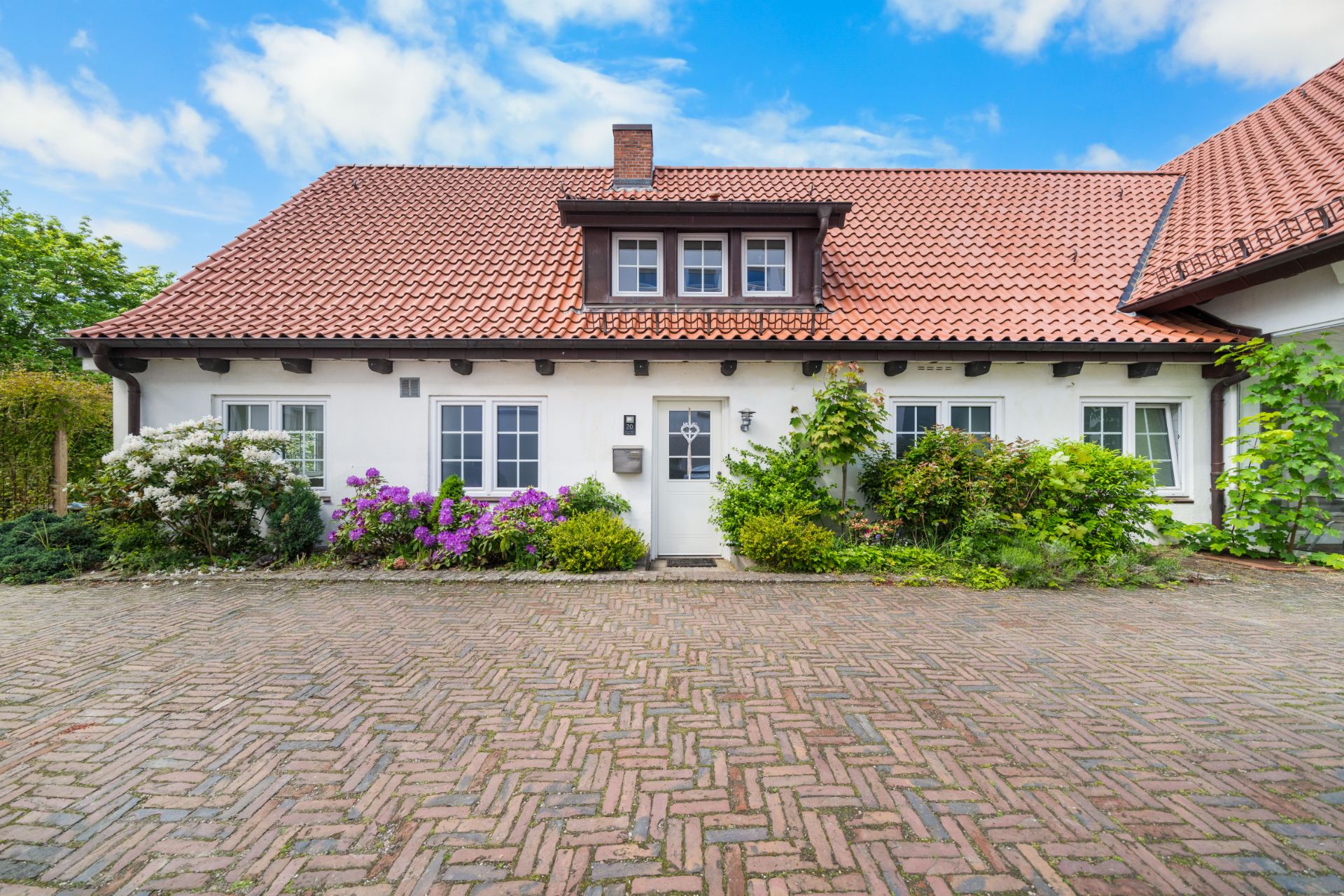 Haus mit rotem Ziegeldach, weißen Wänden und Blumenbeeten vor dem Eingang.