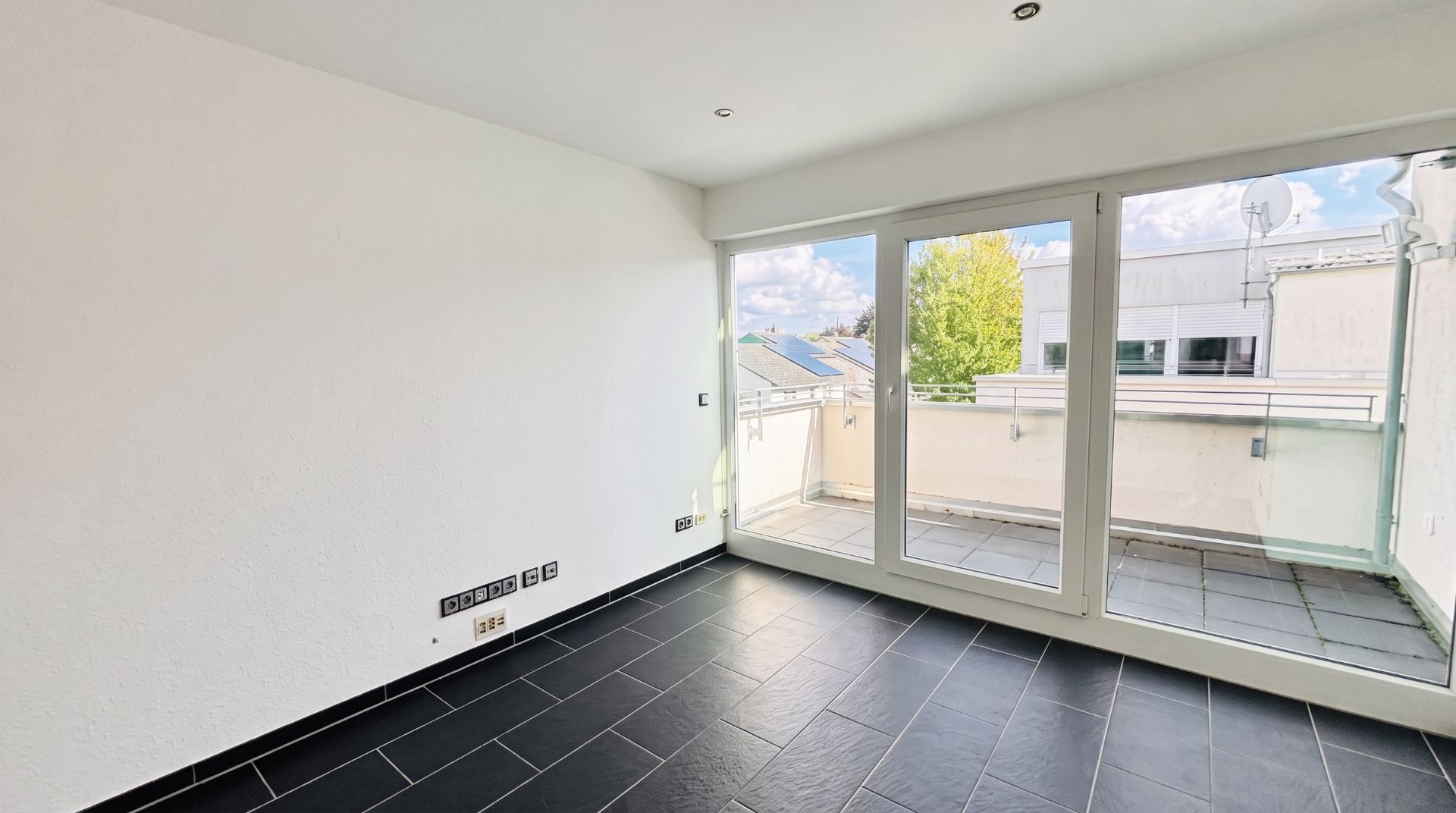 Heller Raum mit dunklem Fliesenboden und Zugang zu einem Balkon durch große Glasfenster.