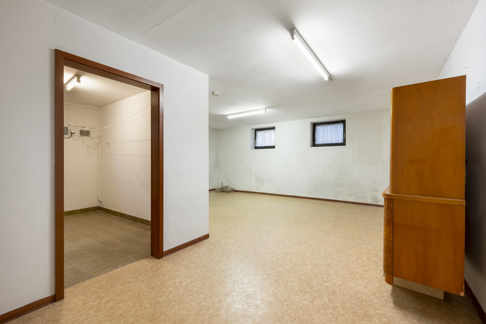 Leerer Kellerraum mit zwei kleinen Fenstern und einem Holzschrank, angrenzender Nebenraum sichtbar.