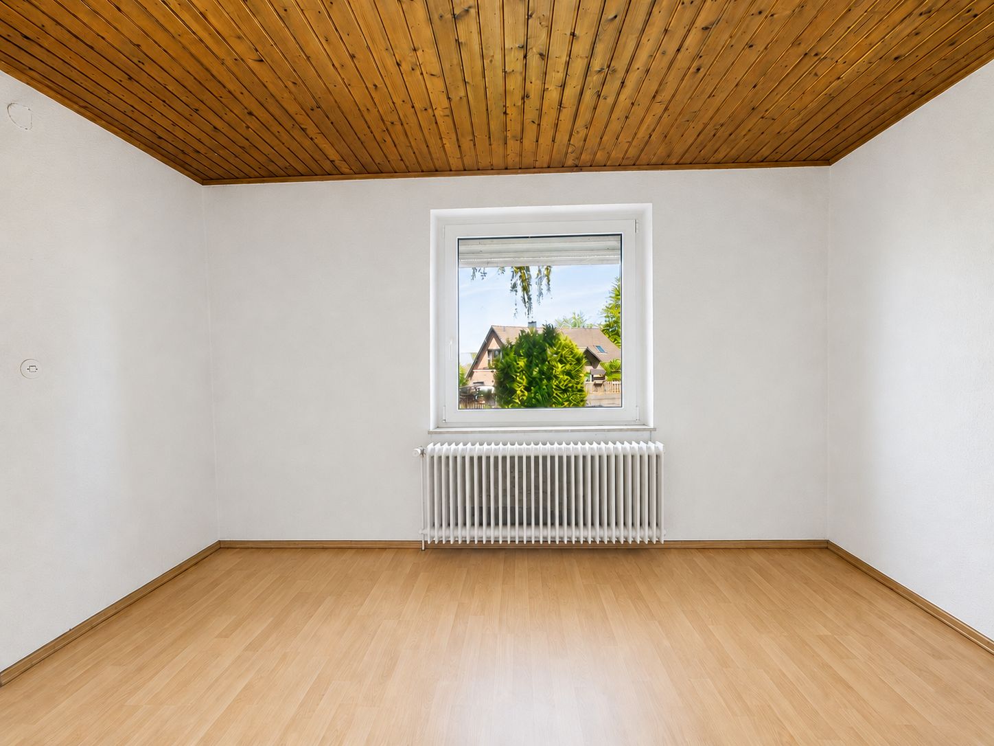 Leerer Raum mit Holzdecke, hellen Wänden, Laminatboden und einem Fenster mit Heizkörper.