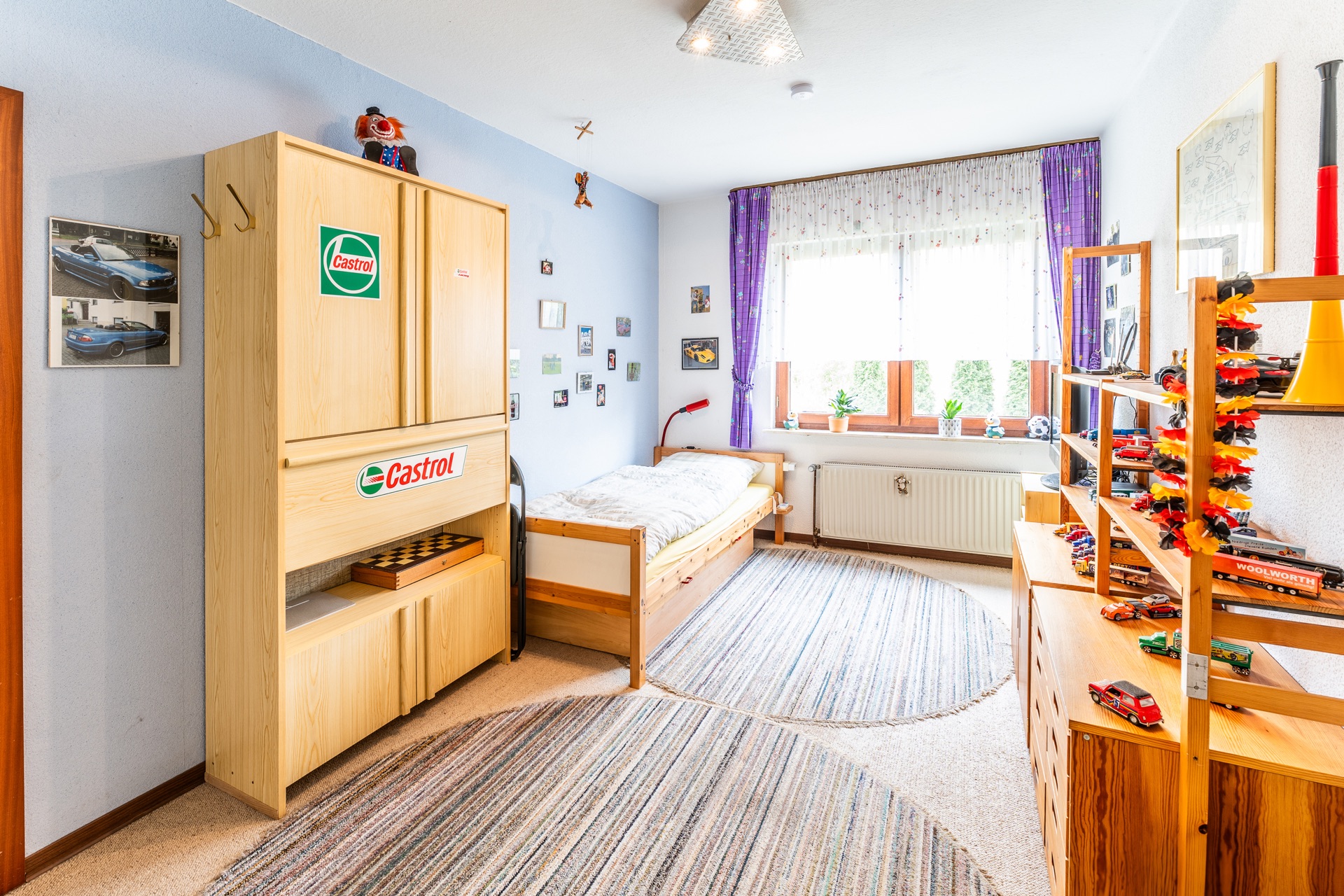Helles Kinderzimmer mit Einzelbett, Holzschrank, Spielzeugautos und großem Fenster mit lila Vorhängen.