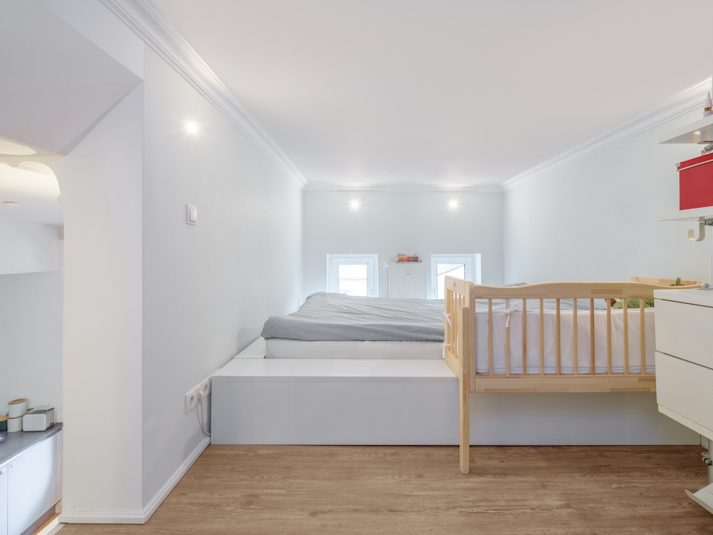 Helles Schlafzimmer mit Doppelbett und Babybett auf erhöhtem Podest, Holzboden, weiß gestrichene Wände.