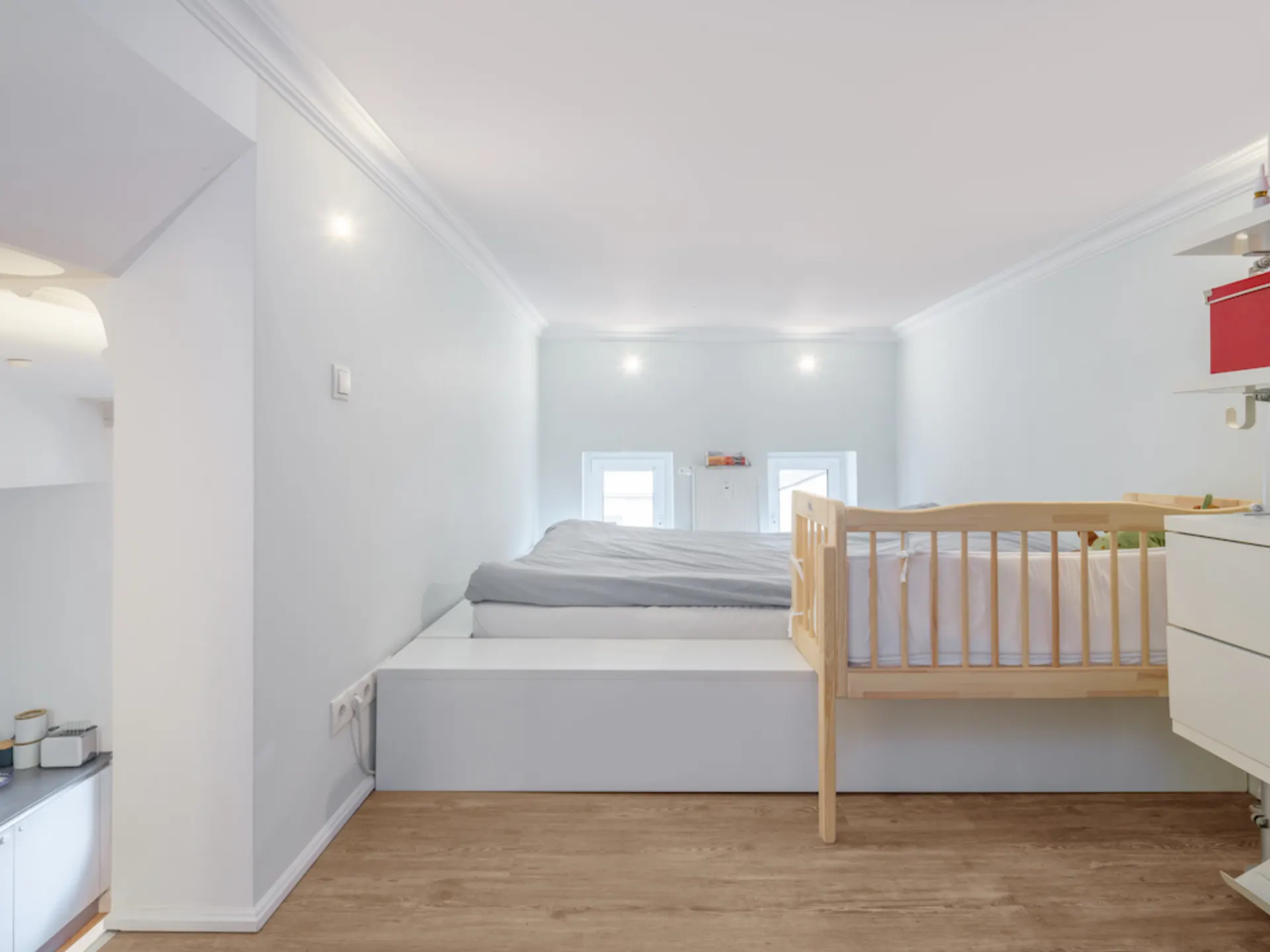Helles Schlafzimmer mit Doppelbett und Babybett auf erhöhtem Podest, Holzboden, weiß gestrichene Wände.