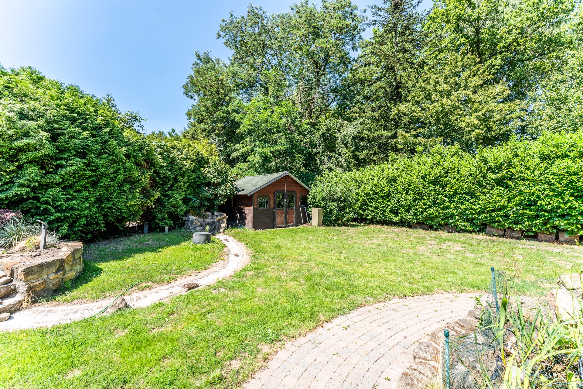 Garten mit Rasenfläche, Gartenhaus und umliegenden Bäumen und Sträuchern bei Sonnenschein.