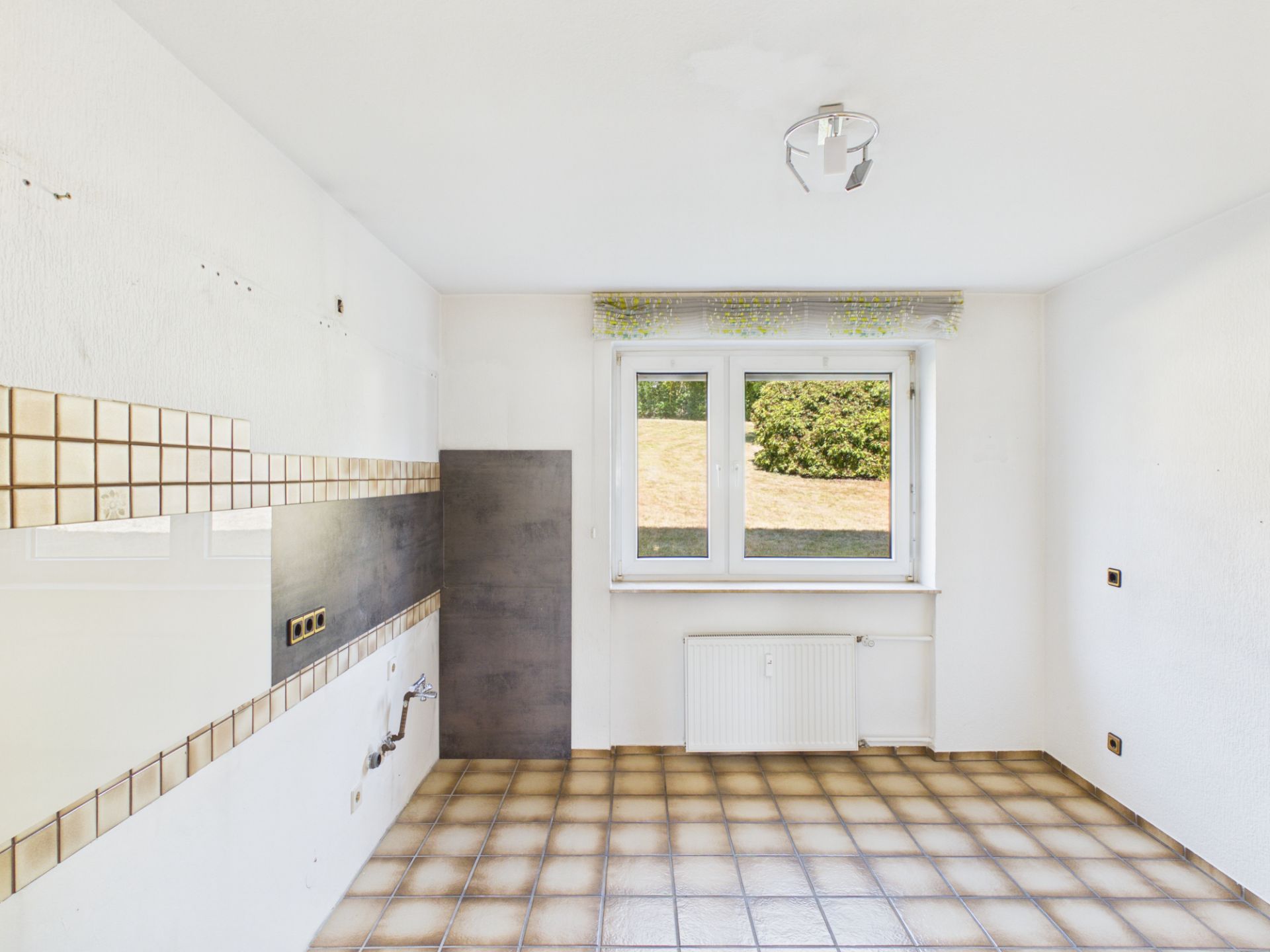 Leerer Raum mit gefliestem Boden, Fenster, Heizung und Wandanschlüssen für eine Küche.