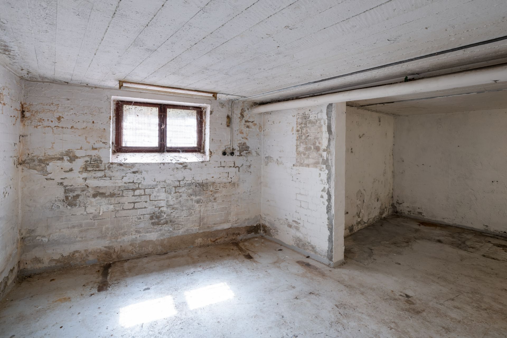 Leerer, alter Kellerraum mit weiß gestrichenen Wänden und kleinem Fenster.