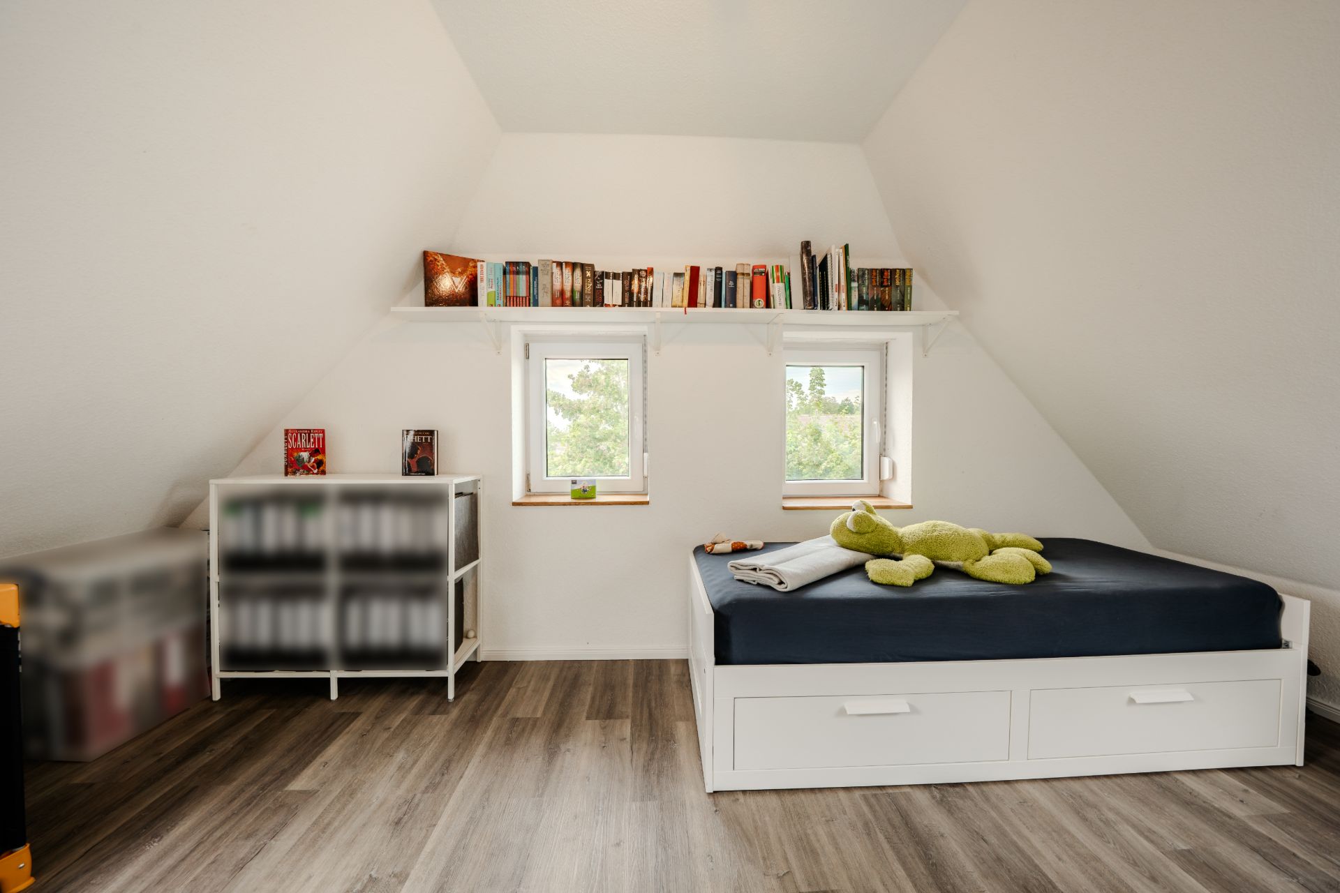 Helles Schlafzimmer mit Bett, Regal, zwei Fenstern und Bücherregal an der Wand.